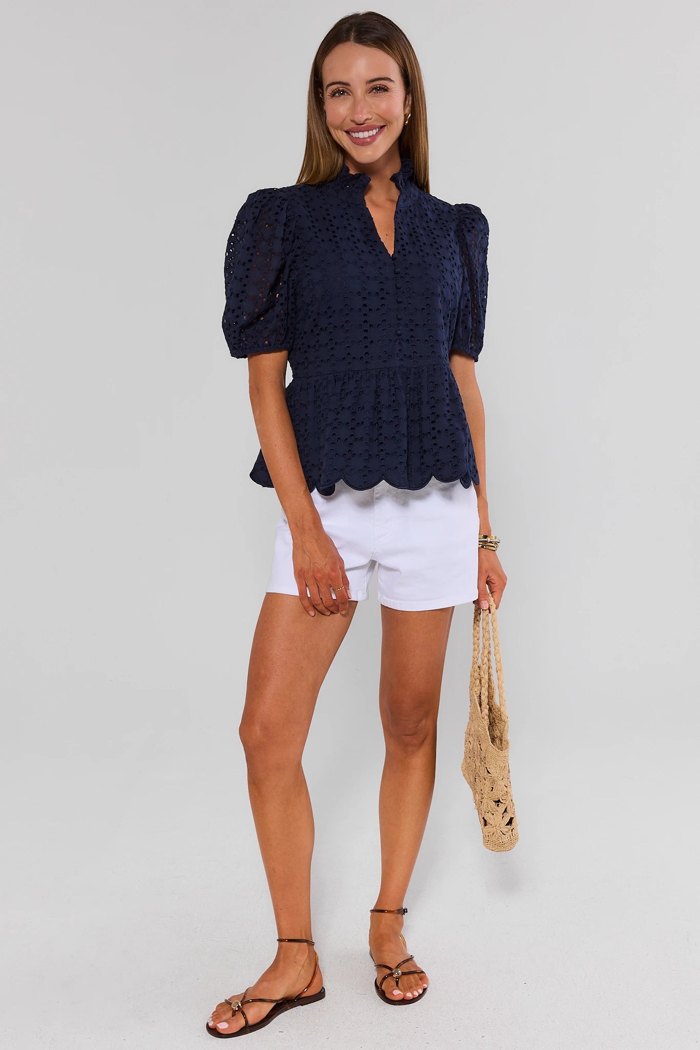 Woman wearing a navy blue top and white shorts, holding a straw bag on a plain background standing