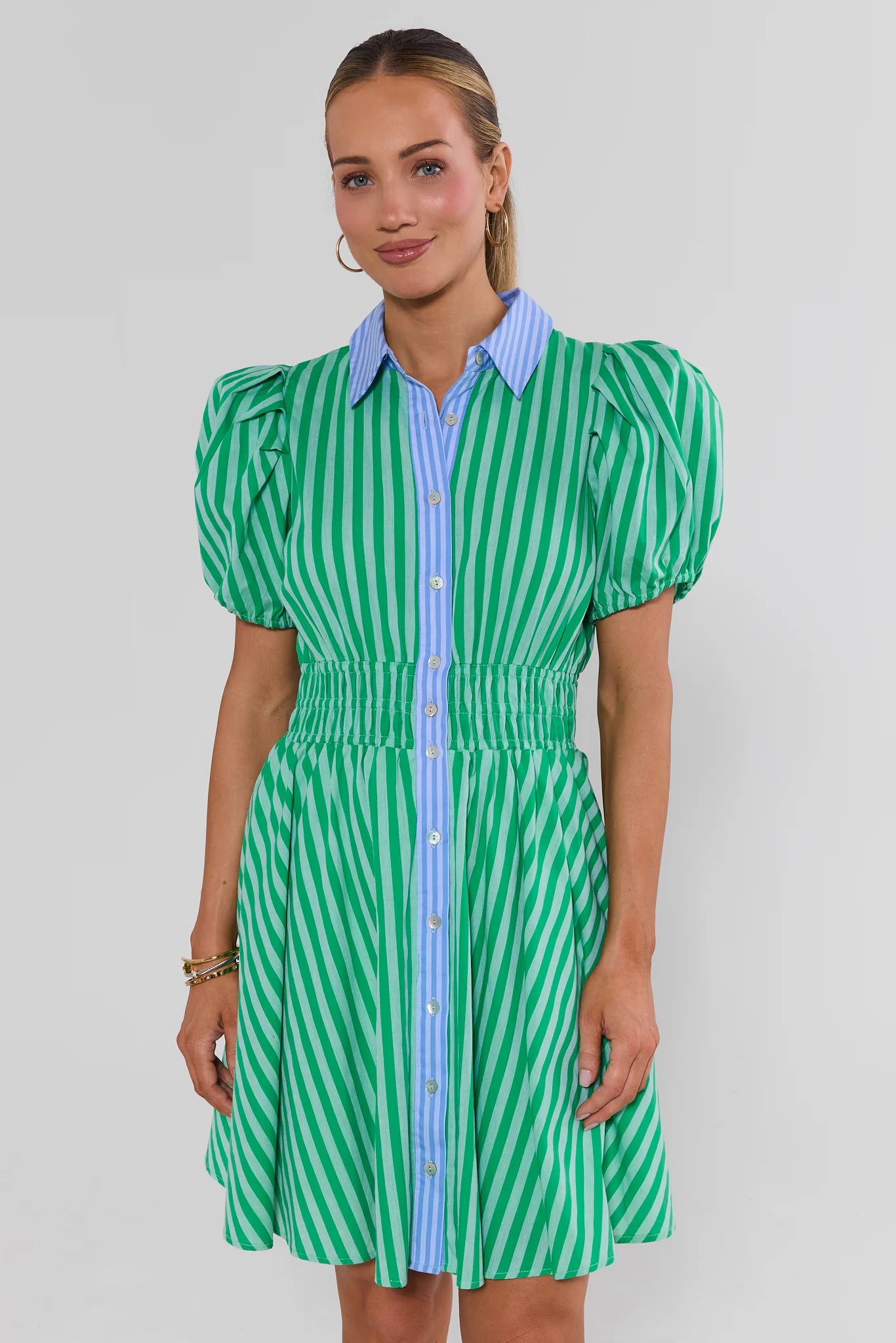 Woman wearing a green and white striped dress with a blue shirt underneath on a gray background standing