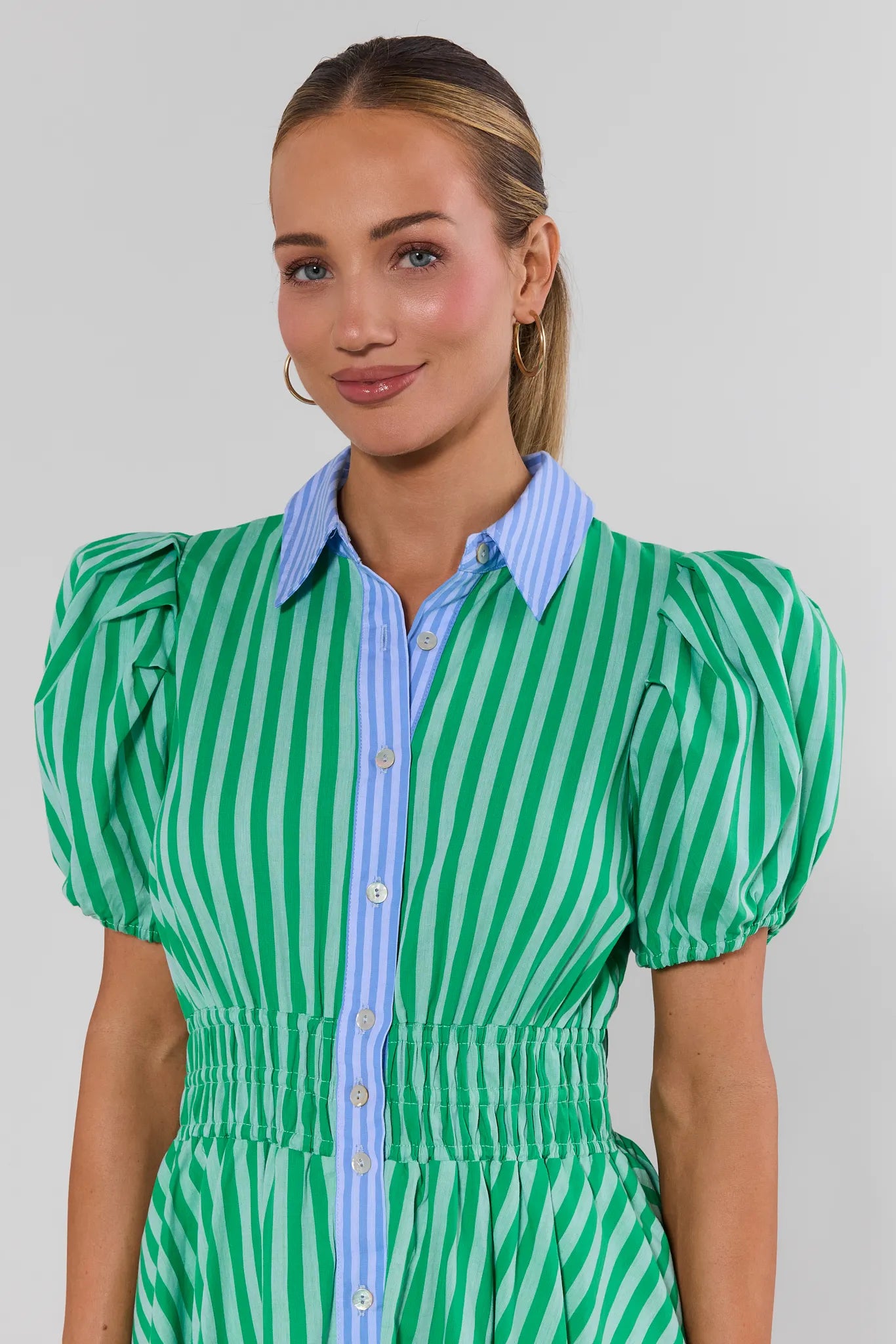 Woman wearing a green and white striped dress with a blue shirt underneath on a plain background close up