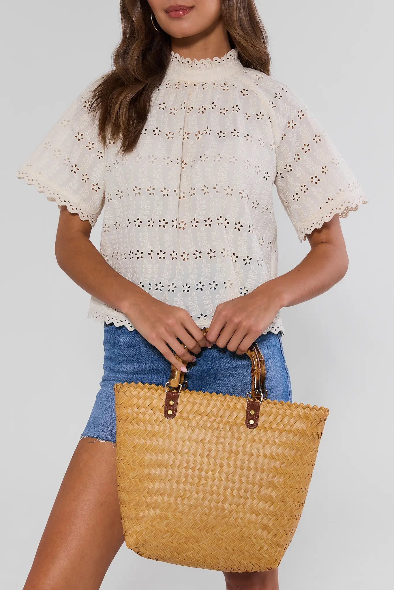 Woman holding a straw bag wearing a white lace top and blue shorts on a plain background