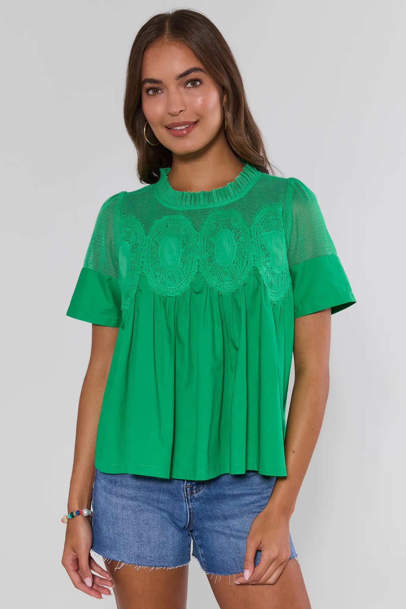 Woman wearing a green blouse with a textured pattern and denim shorts on a plain background