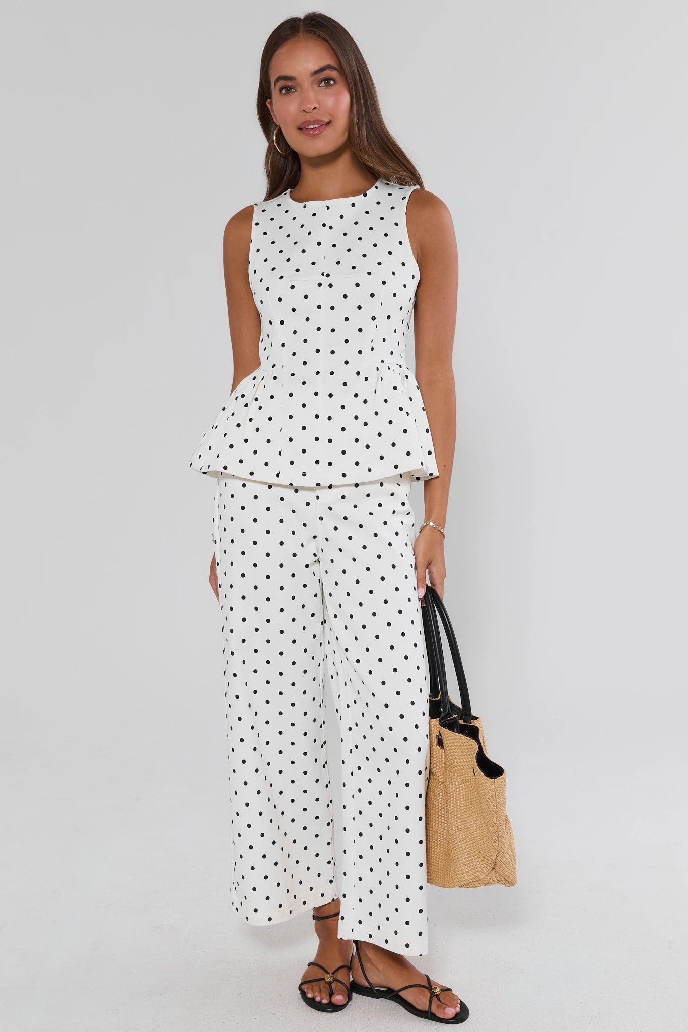Woman wearing a white sleeveless top and pants with black polka dots, holding a beige bag on a light gray background.