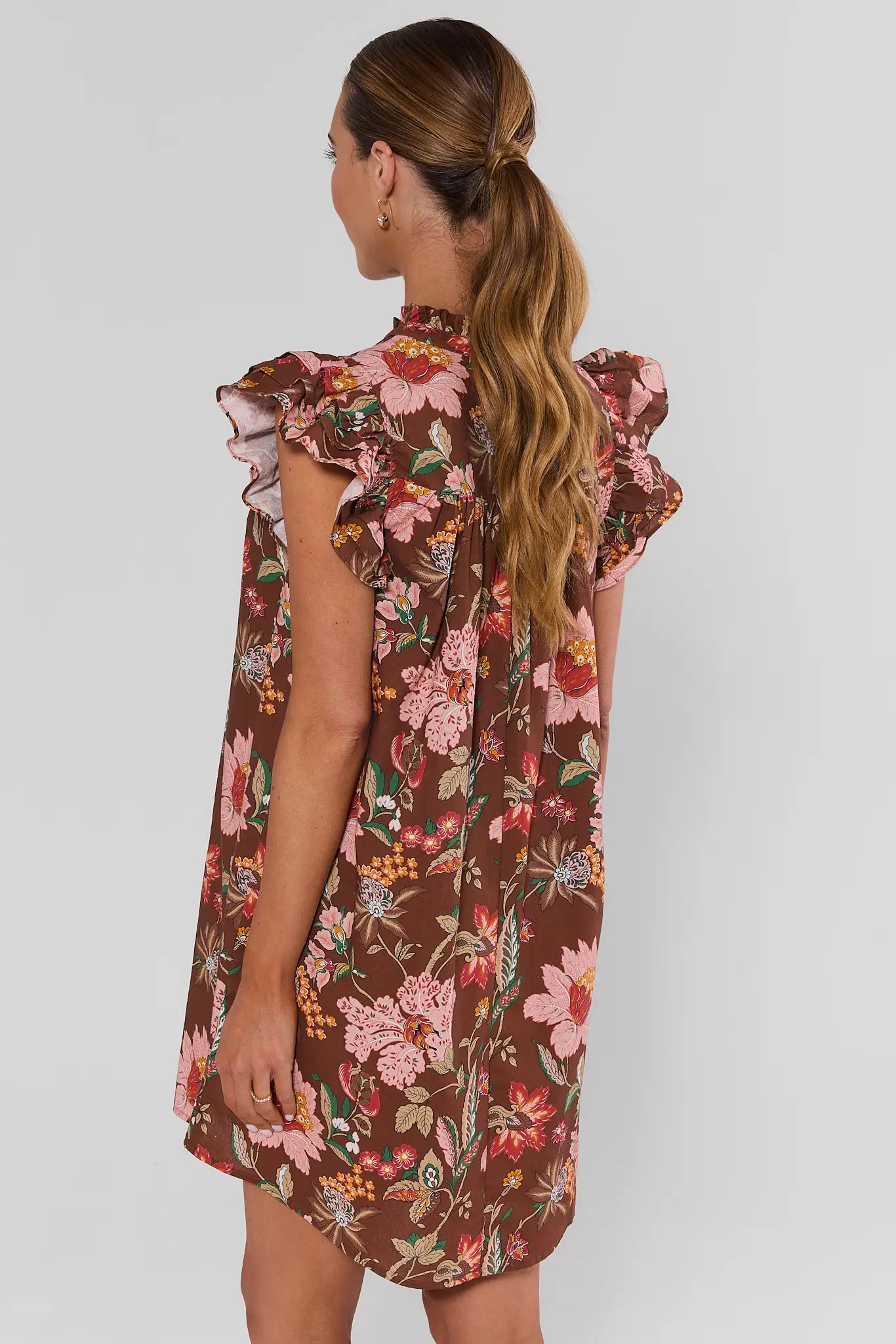 A woman modeling a brown short-sleeve floral dress with ruffled shoulders and a buttoned collar, on a plain background.