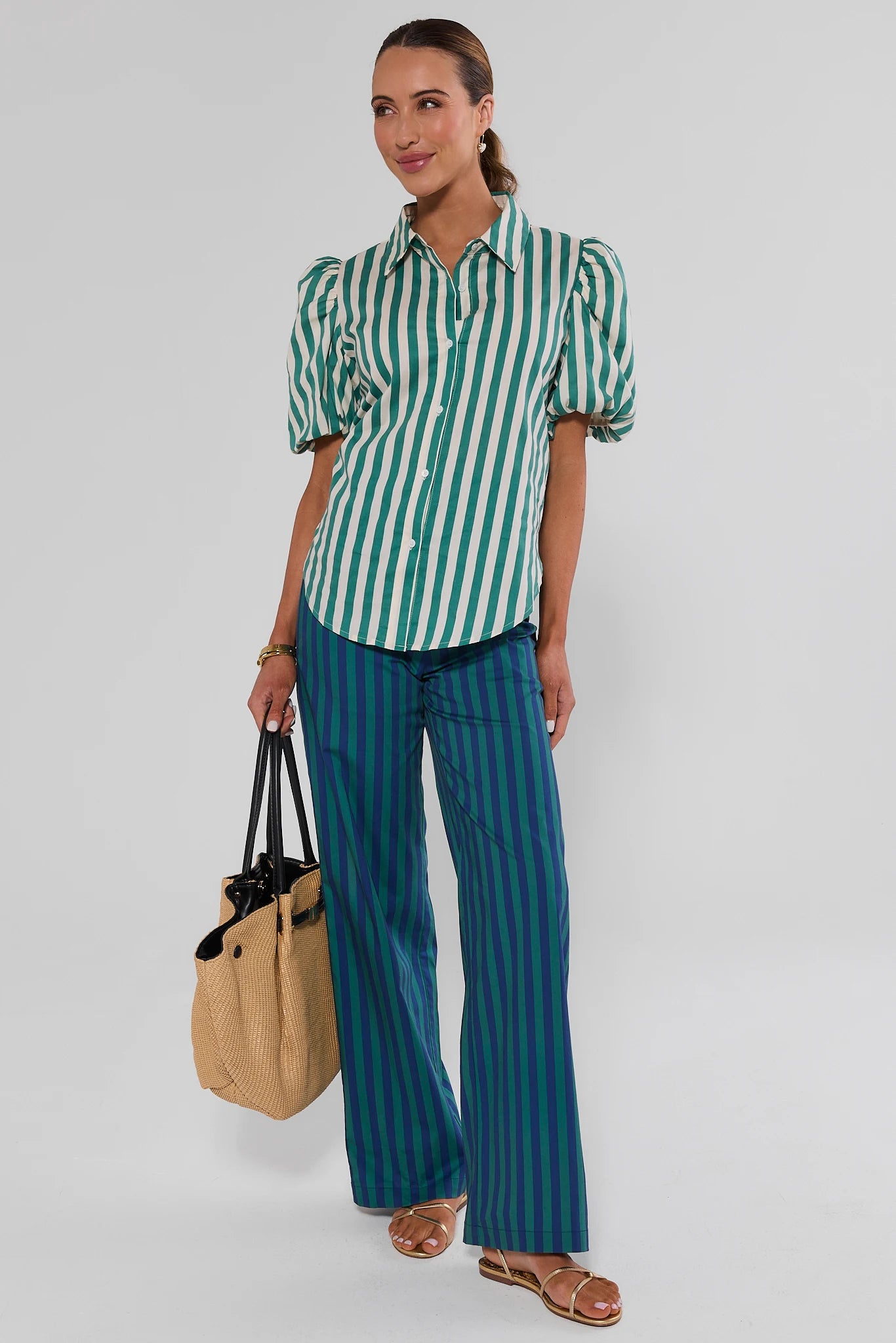 A woman standing wearing a green and white vertically striped crop top with puff sleeves and a tie-back waist, paired with blue and green striped pants, on a white background