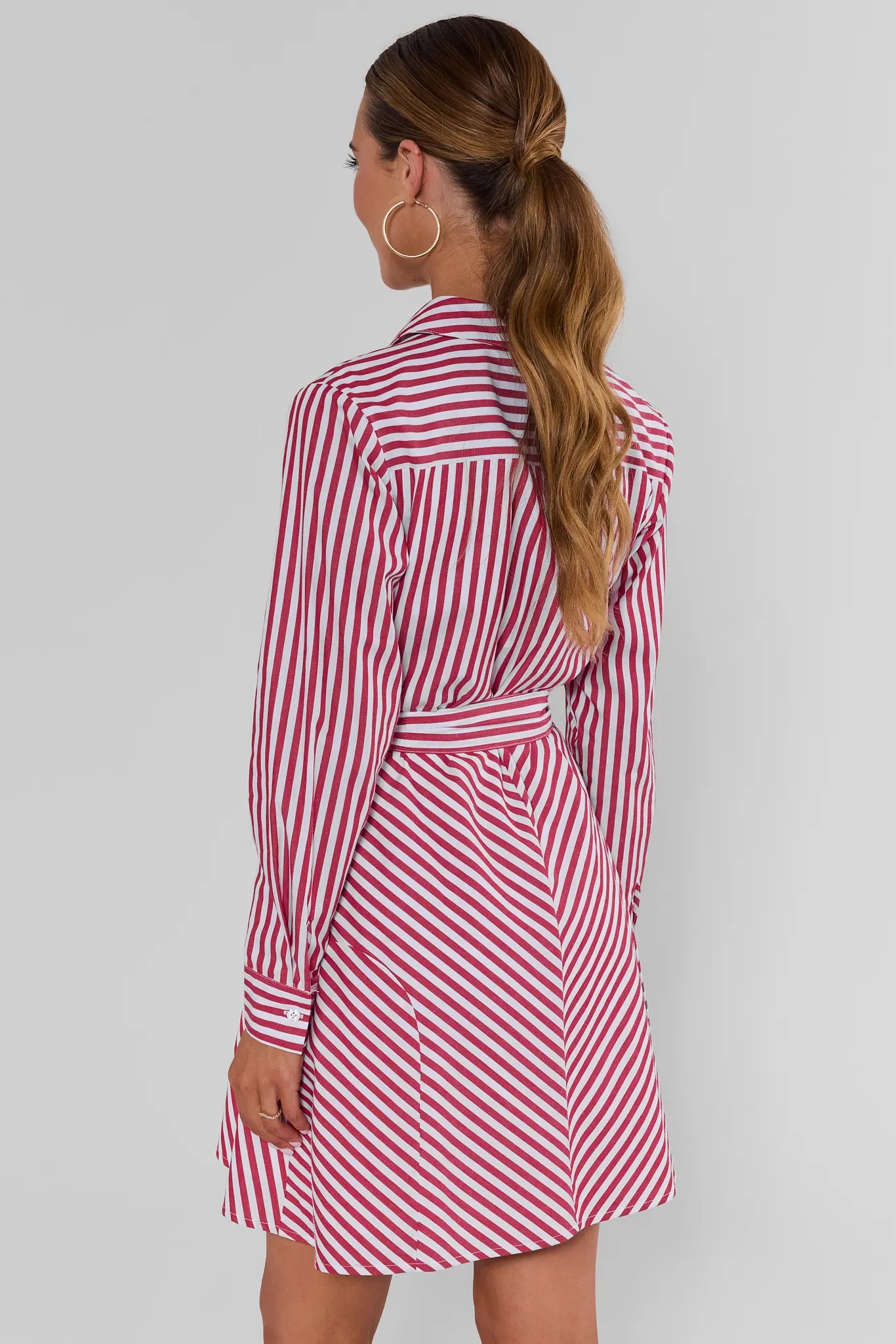 A woman turned wearing a red and white vertically striped shirt dress with a tie waist, photographed against a light gray background.