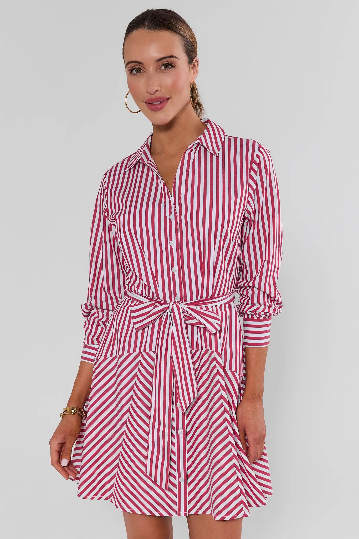 A woman standing wearing a red and white vertically striped shirt dress with a tie waist, photographed against a light gray background.