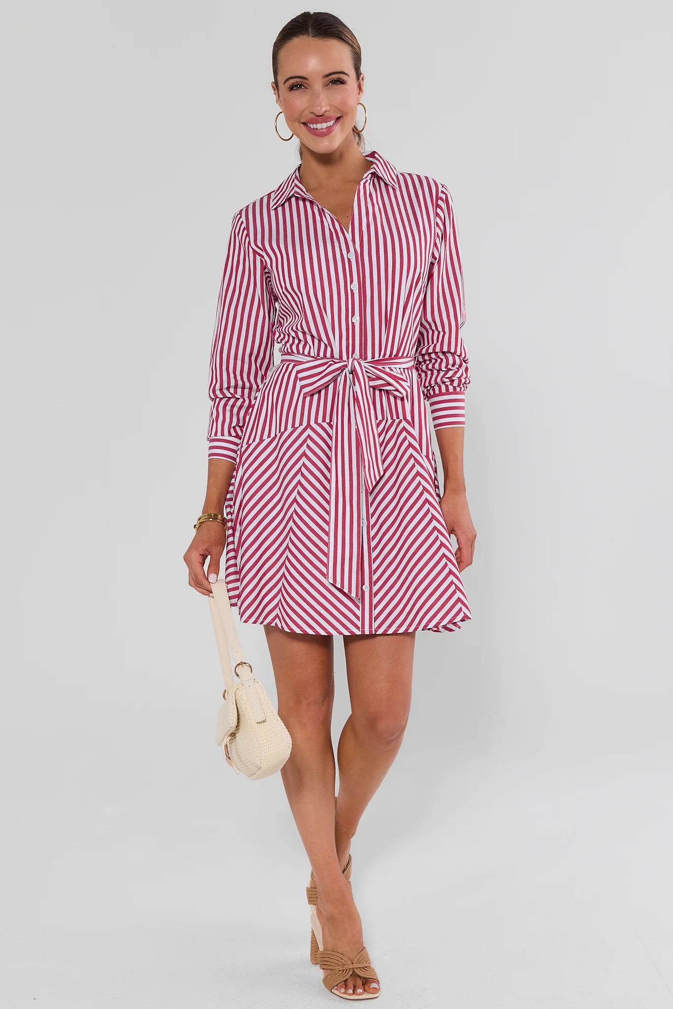 A woman modeling a pink and white striped shirt dress with a tie waist, holding a cream-colored handbag against a light gray studio background.