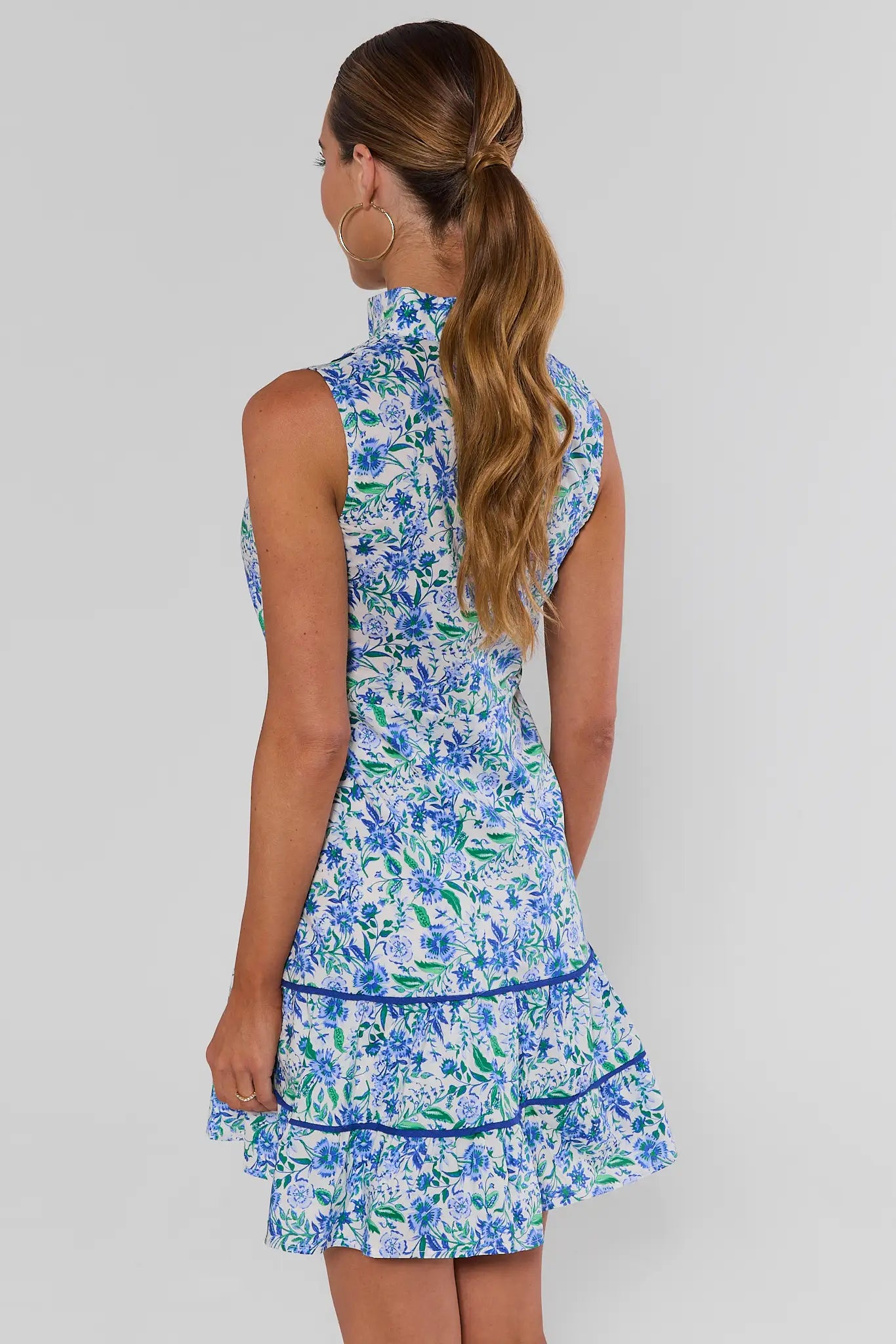 A woman standing backwards wearing a sleeveless high-neck mini dress with a blue and green floral print on a white background, set against a plain light gray studio background.