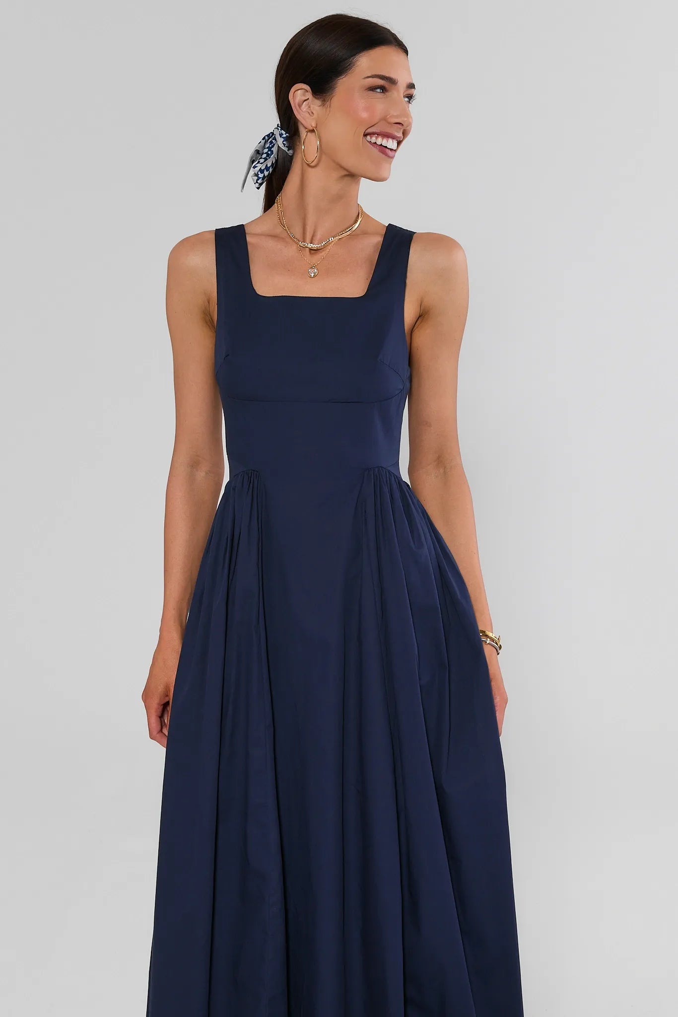 A woman modeling a sleeveless navy blue maxi dress with a square neckline and a flared skirt, photographed against a plain light gray studio background.
