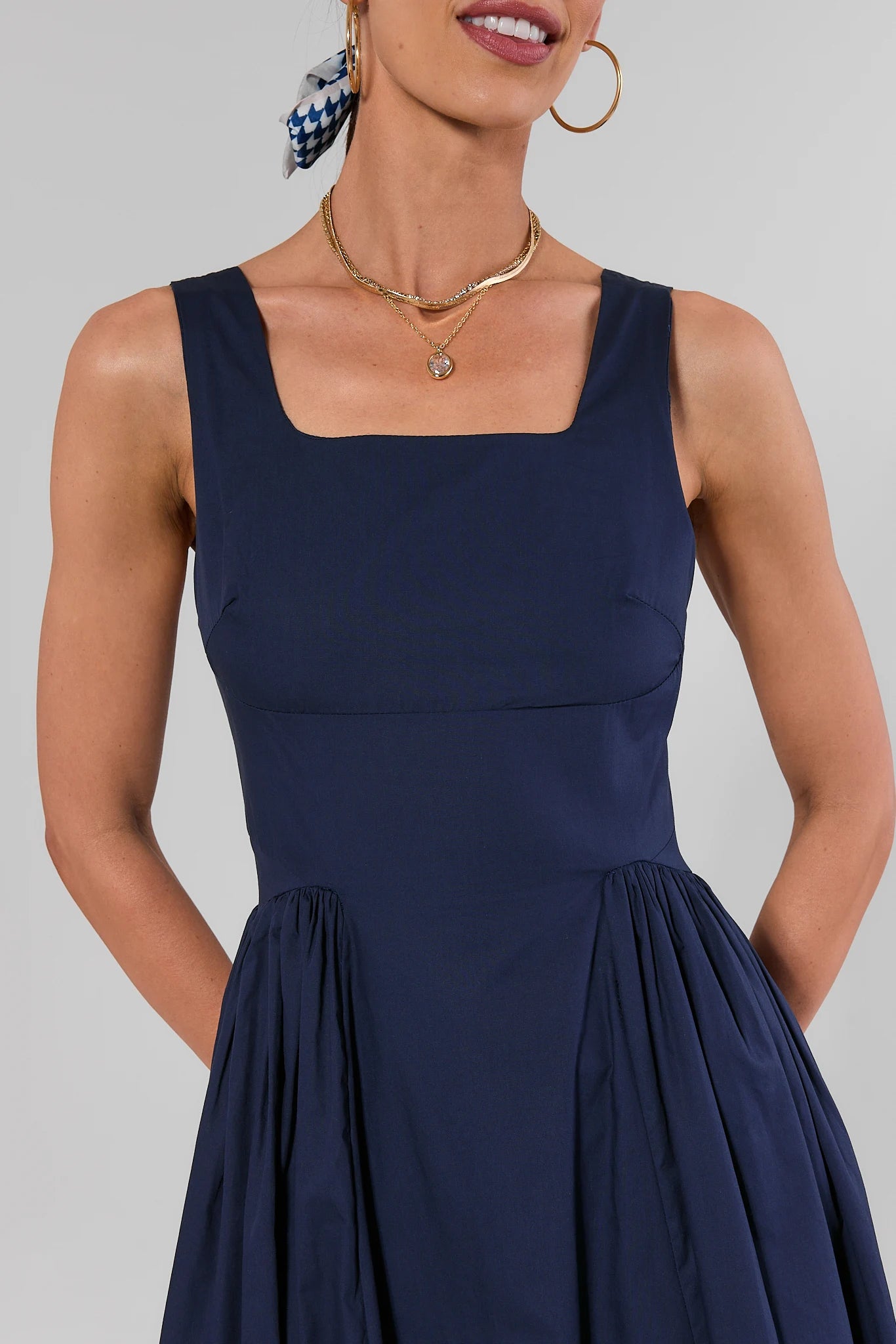 A woman wearing a navy blue sleeveless dress with a square neckline, structured bodice, and gathered skirt, shown against a plain light gray studio background.