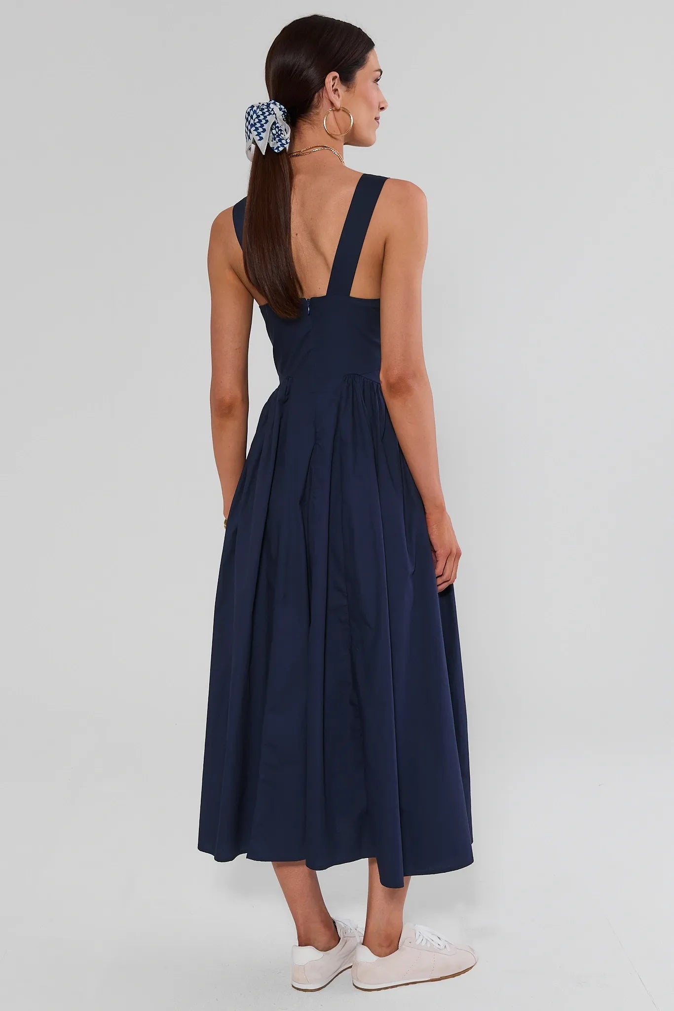 A woman modeling a navy blue midi dress from the back, featuring a square neckline, wide shoulder straps, a gathered waist, and a flared skirt. She is wearing white sneakers and a blue-and-white patterned scrunchie against a gray background.