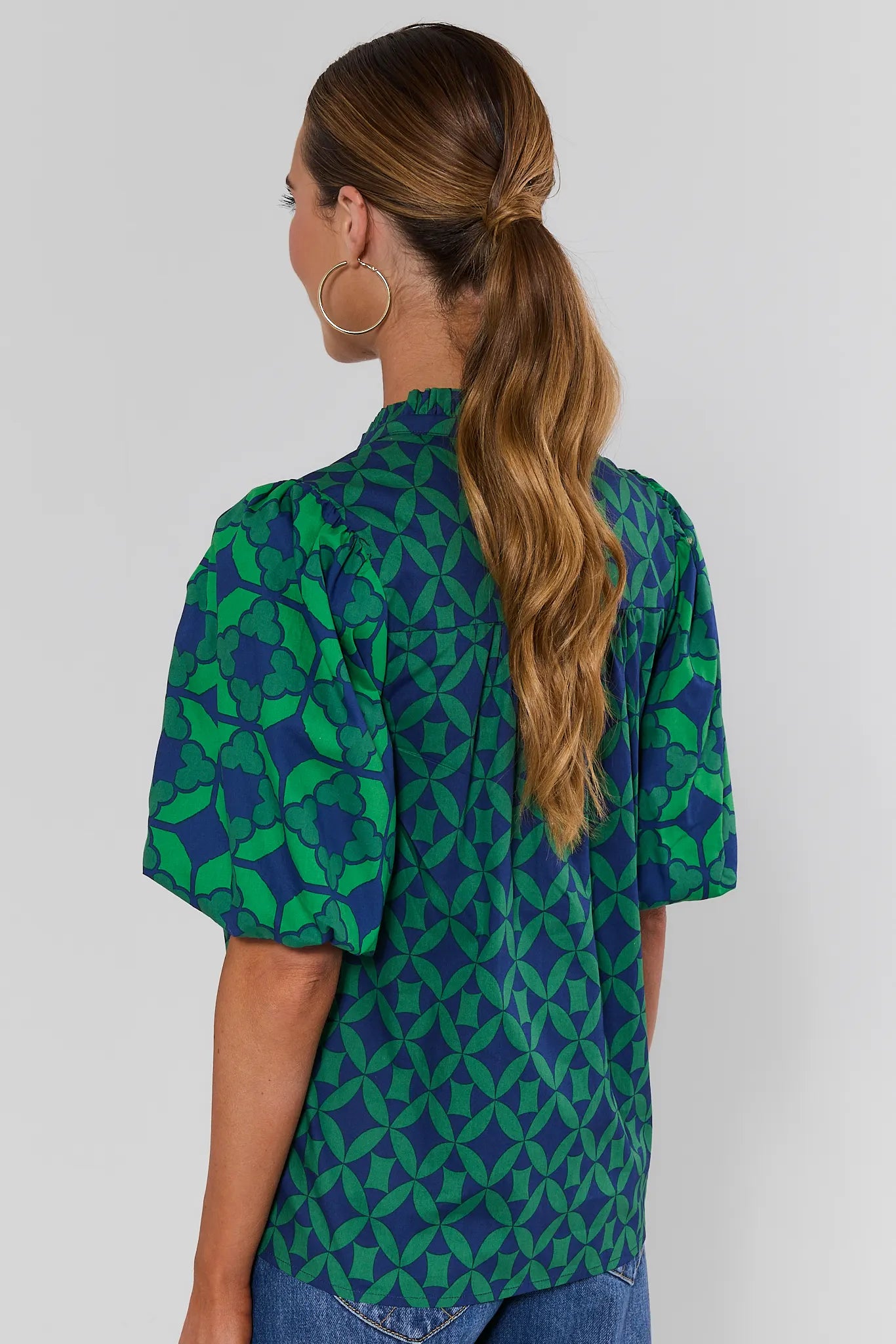 A woman turned backwards wearing a green and blue patterned short-sleeve blouse with puff sleeves against a white background