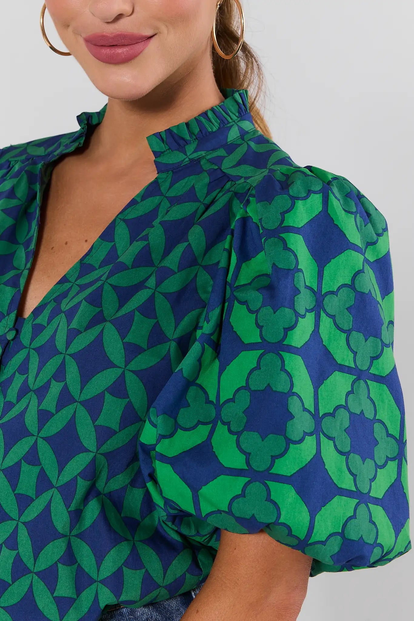 A close-up of a woman wearing a short-sleeve blouse with a bold green and blue geometric pattern and puffed shoulders, paired with gold hoop earrings against a light gray background.