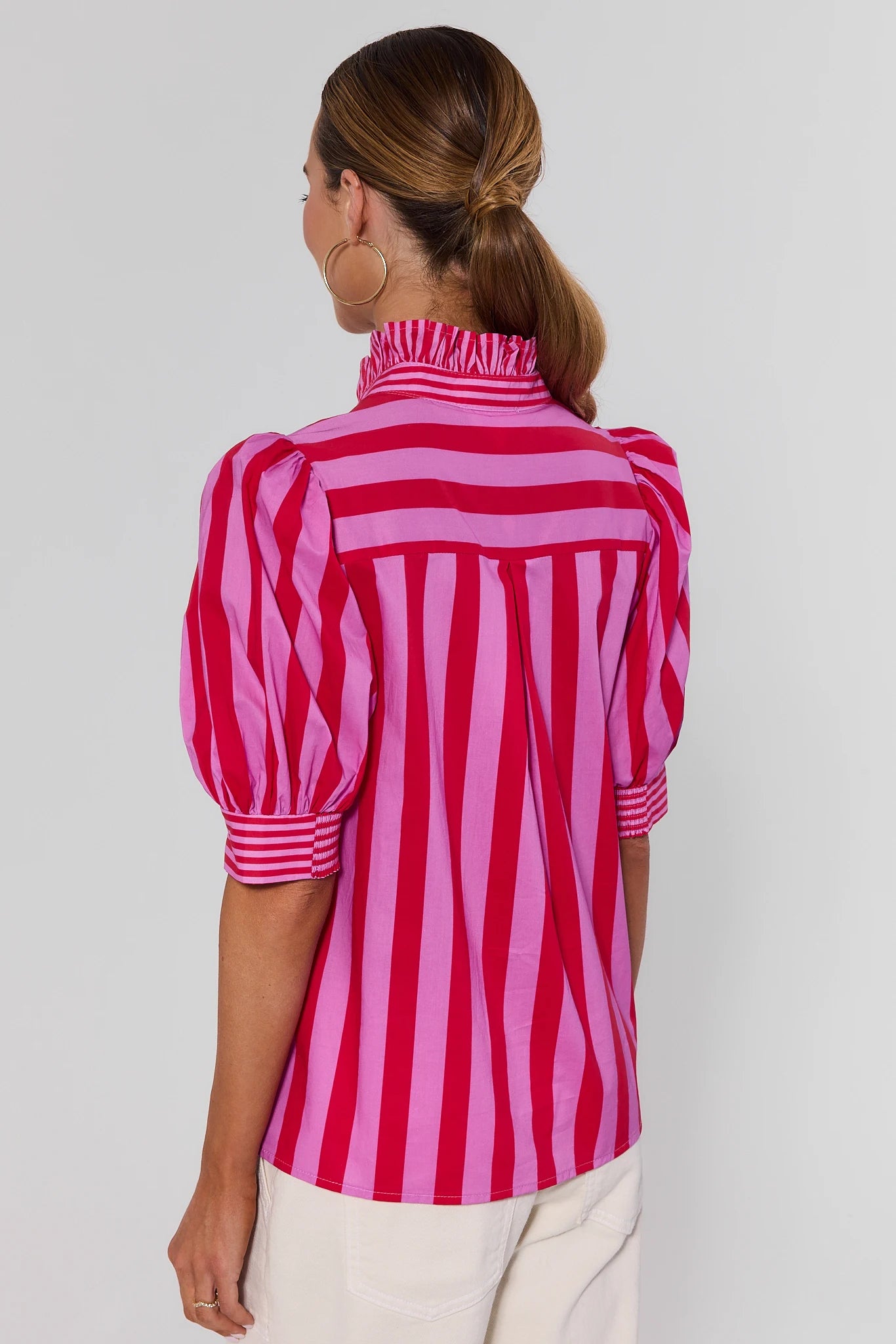 A woman shown from the back wearing a pink and red vertically striped blouse with puff sleeves and a ruffled high neck, paired with off-white pants, against a light gray studio background.