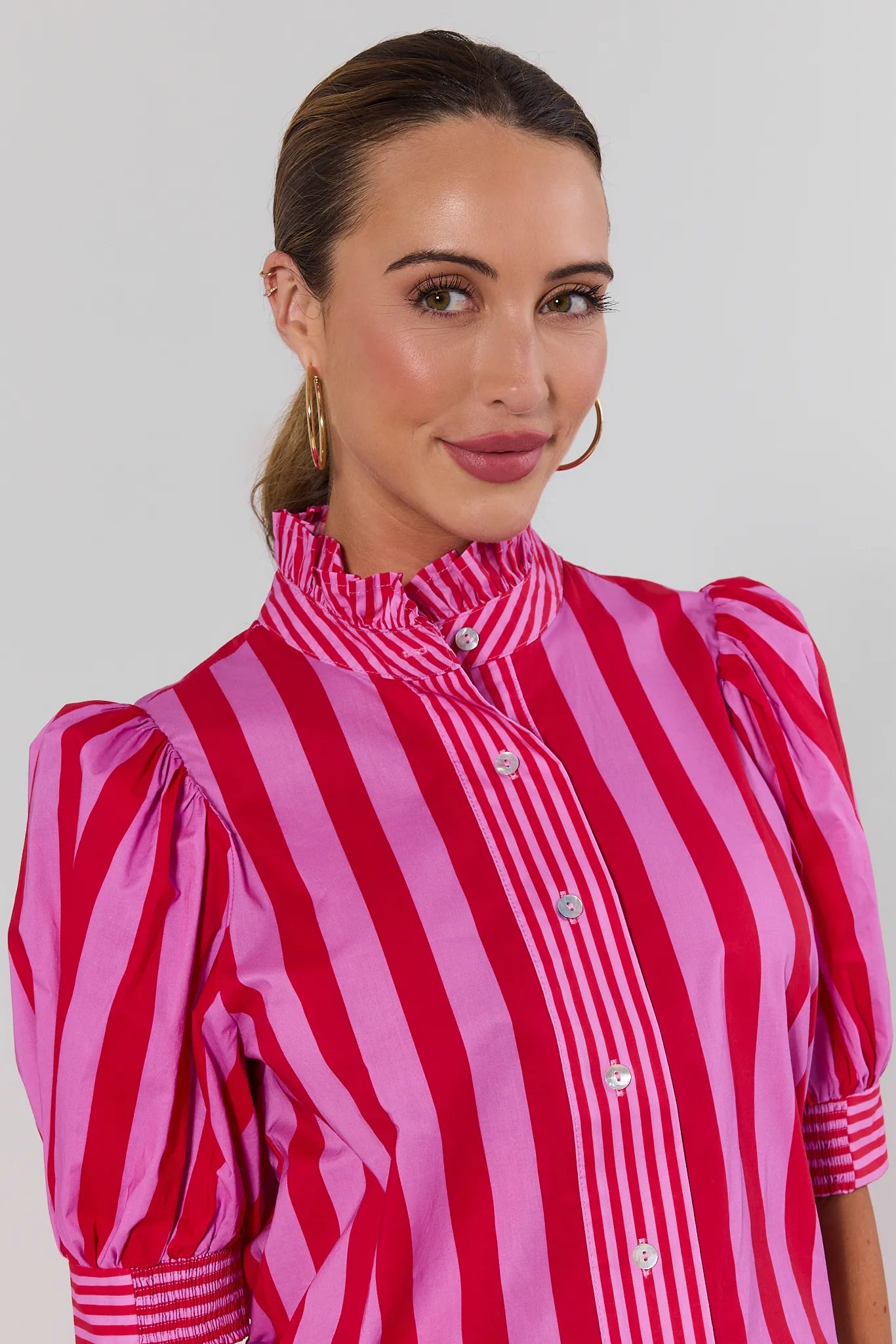 A woman wearing a vibrant pink and red vertically striped blouse with puff sleeves and a high ruffled collar, photographed against a neutral light gray background.