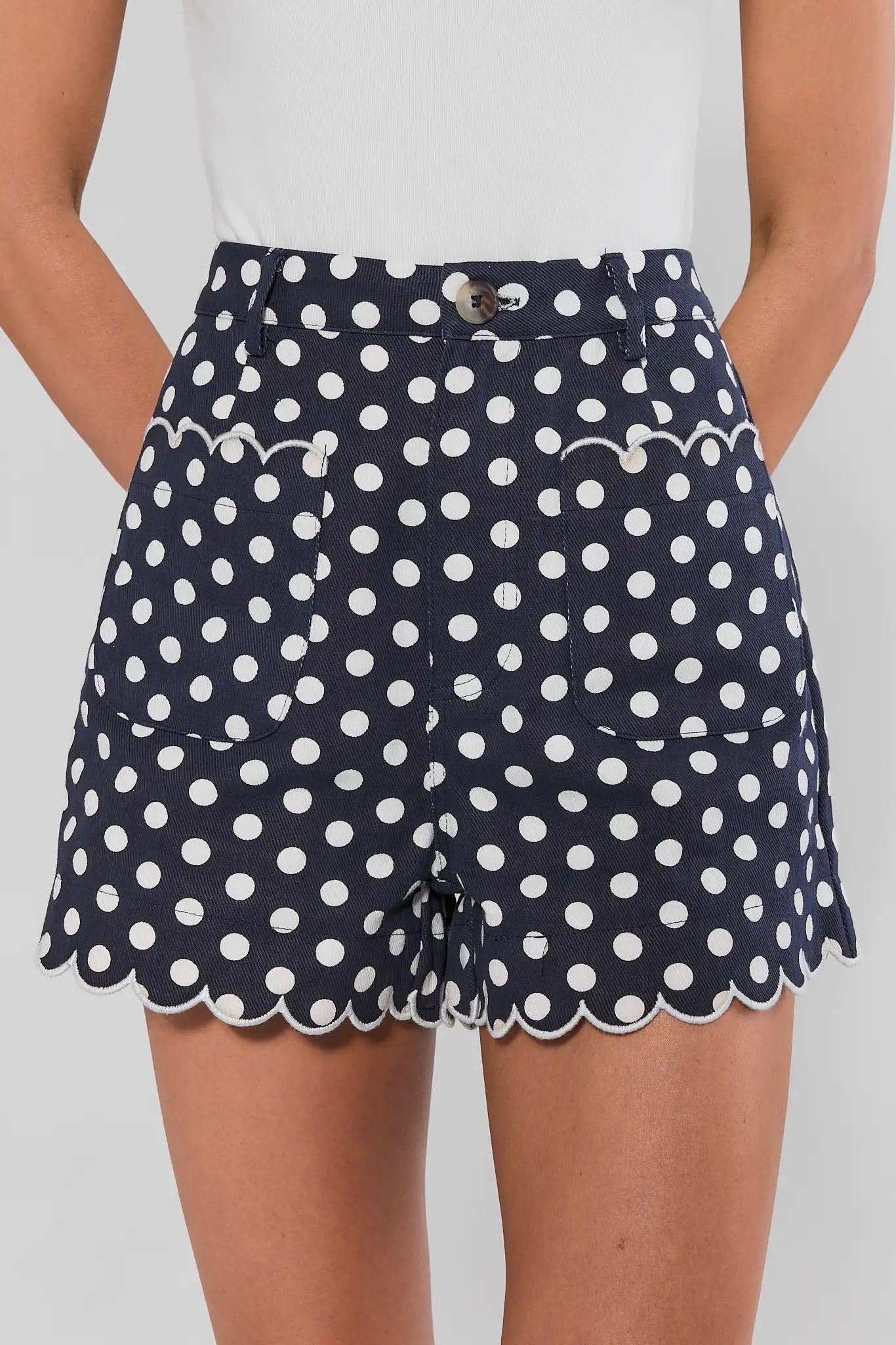 A woman modeling a white top and navy blue shorts with white polka dots and scalloped hems, against a light background.