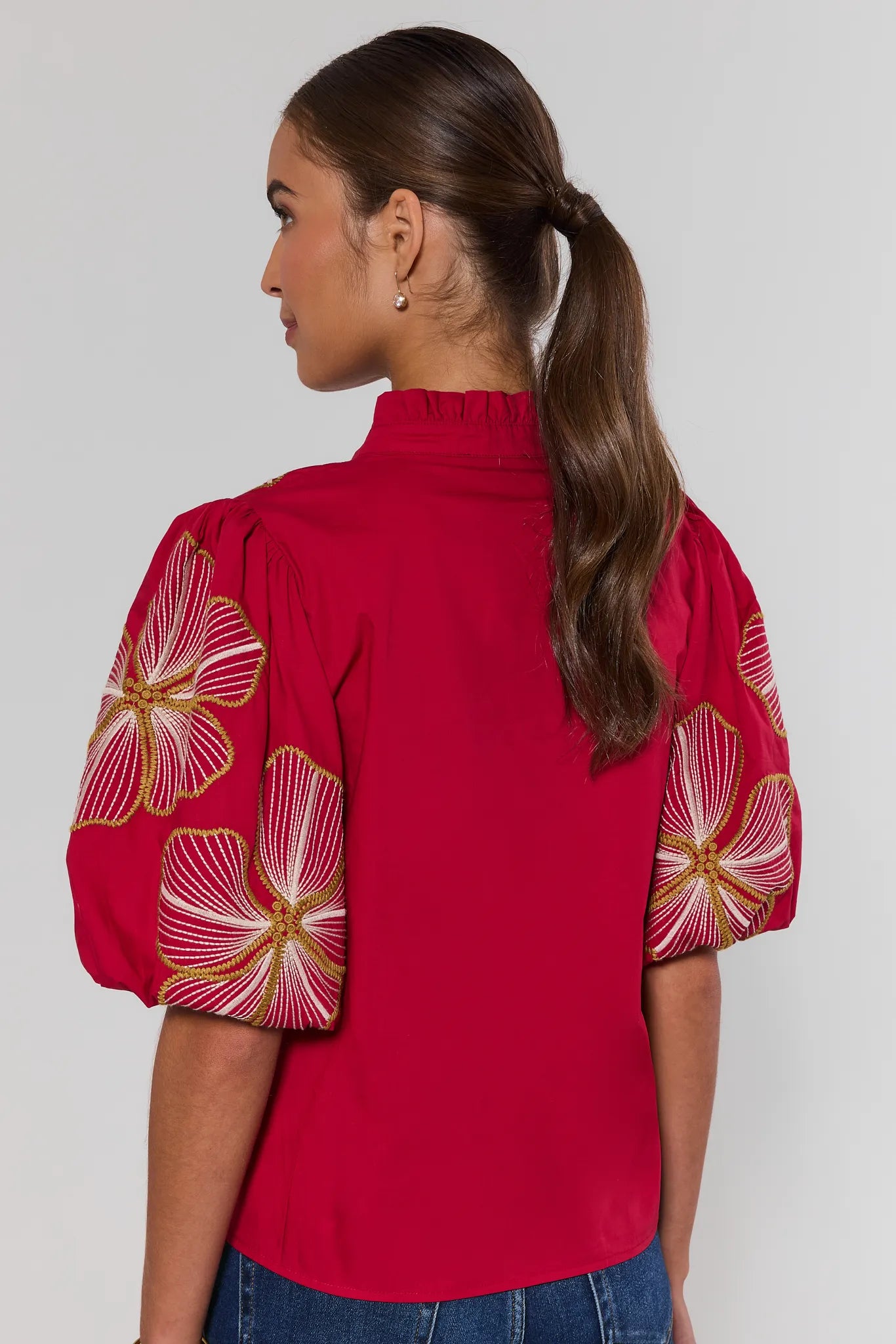 A woman shown from the back wearing a bright red blouse with large embroidered floral details on the sleeves, paired with blue jeans, against a plain light gray studio background.