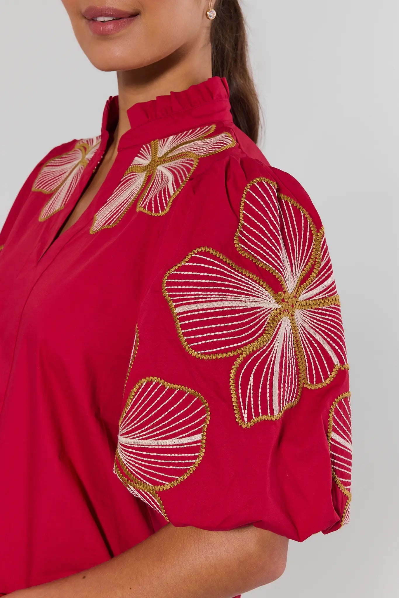 A close-up of a woman wearing a bright red blouse with large white and gold floral embroidery on the bodice and sleeves, paired with a high ruffled collar and puff sleeves.