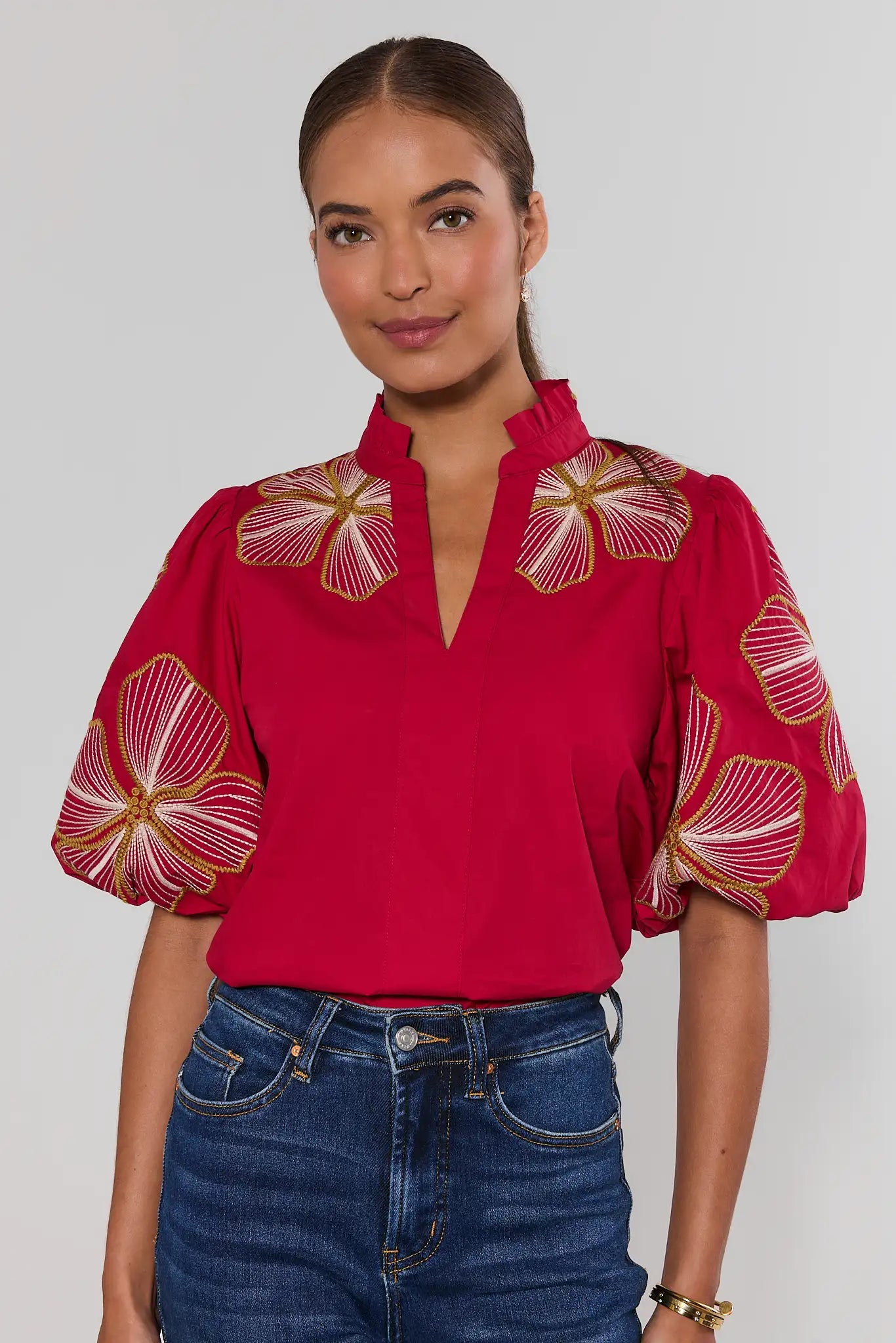 woman wearing a bright red blouse with large white and gold floral embroidery on the bodice and sleeves, paired with a high ruffled collar and puff sleeves.