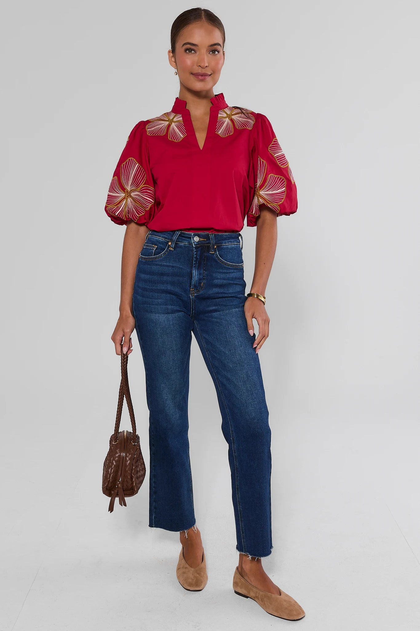 A woman modeling a vibrant red short-sleeve blouse with gold and white floral embroidery on the shoulders and sleeves, paired with high-waisted blue jeans, tan suede loafers, and holding a brown leather handbag against a light gray studio background.