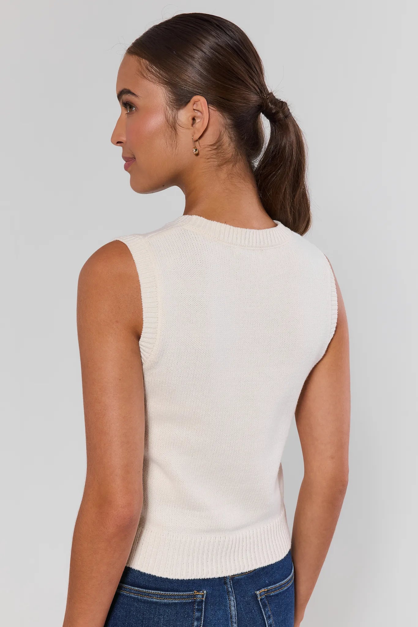 A woman shown from the back wearing a sleeveless cream knit top and blue jeans against a light gray studio background.