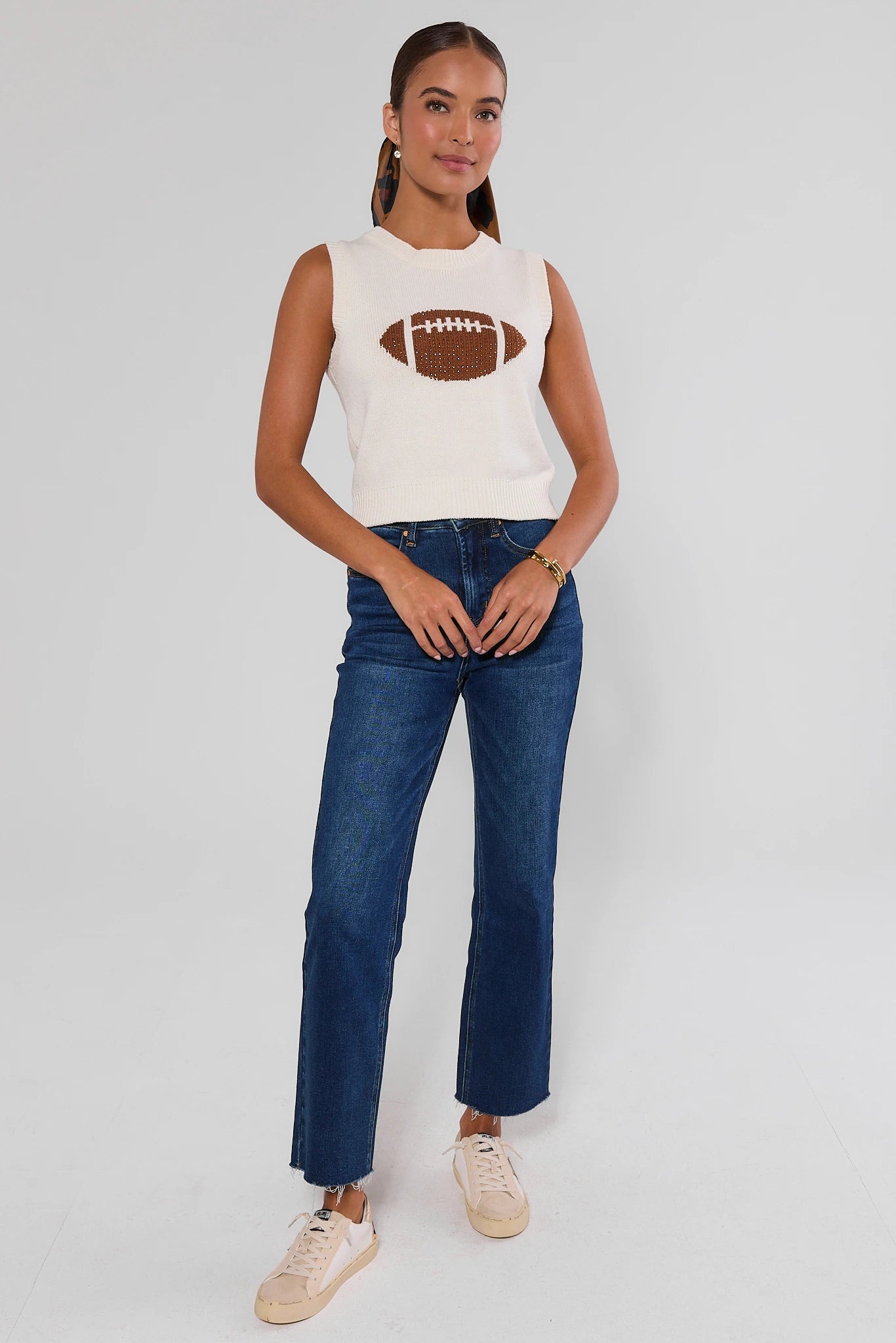 A woman modeling a sleeveless white top with a brown football graphic on the chest, paired with dark blue jeans and beige sneakers, against a light gray studio background.