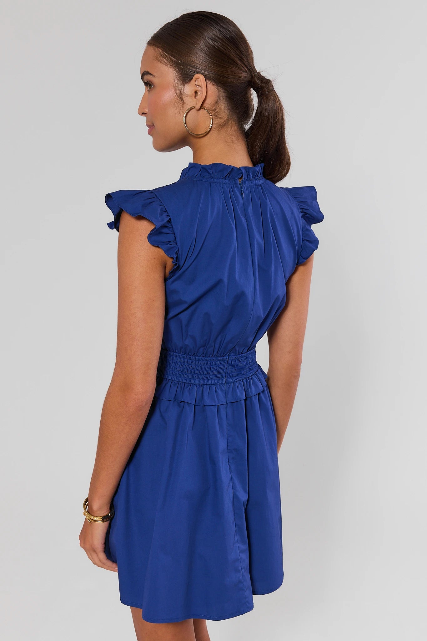 A woman shown from the back wearing a royal blue dress with ruffled cap sleeves and a gathered waist, photographed against a light gray studio background.