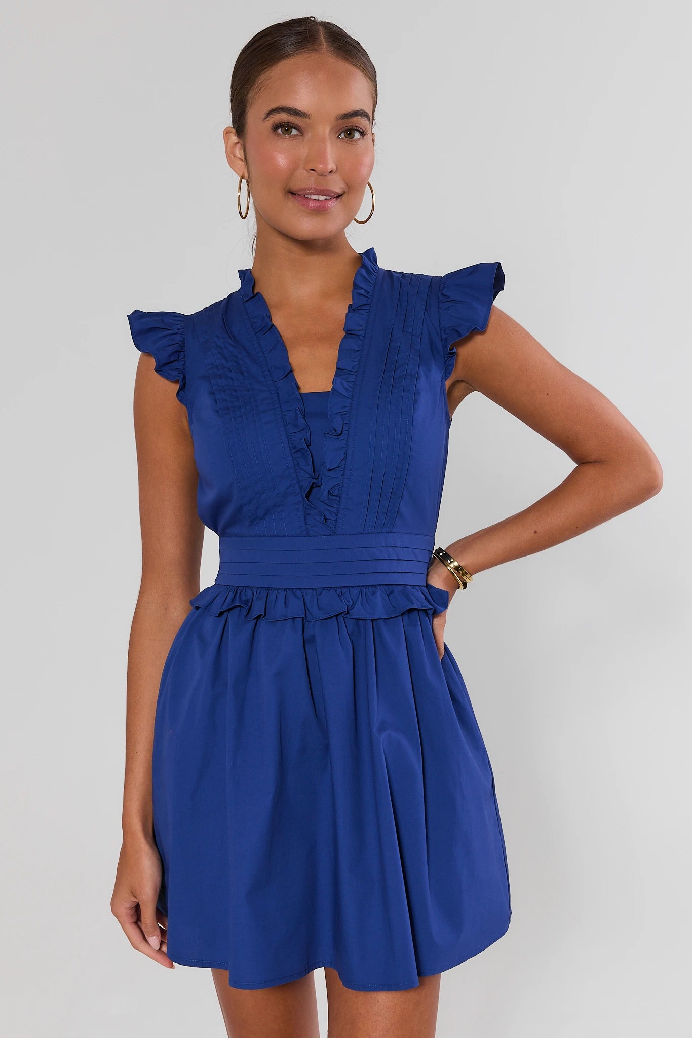 A woman modeling a royal blue sleeveless mini dress with ruffled trim, cap sleeves, a gathered waist, and a flared skirt against a light gray studio background.