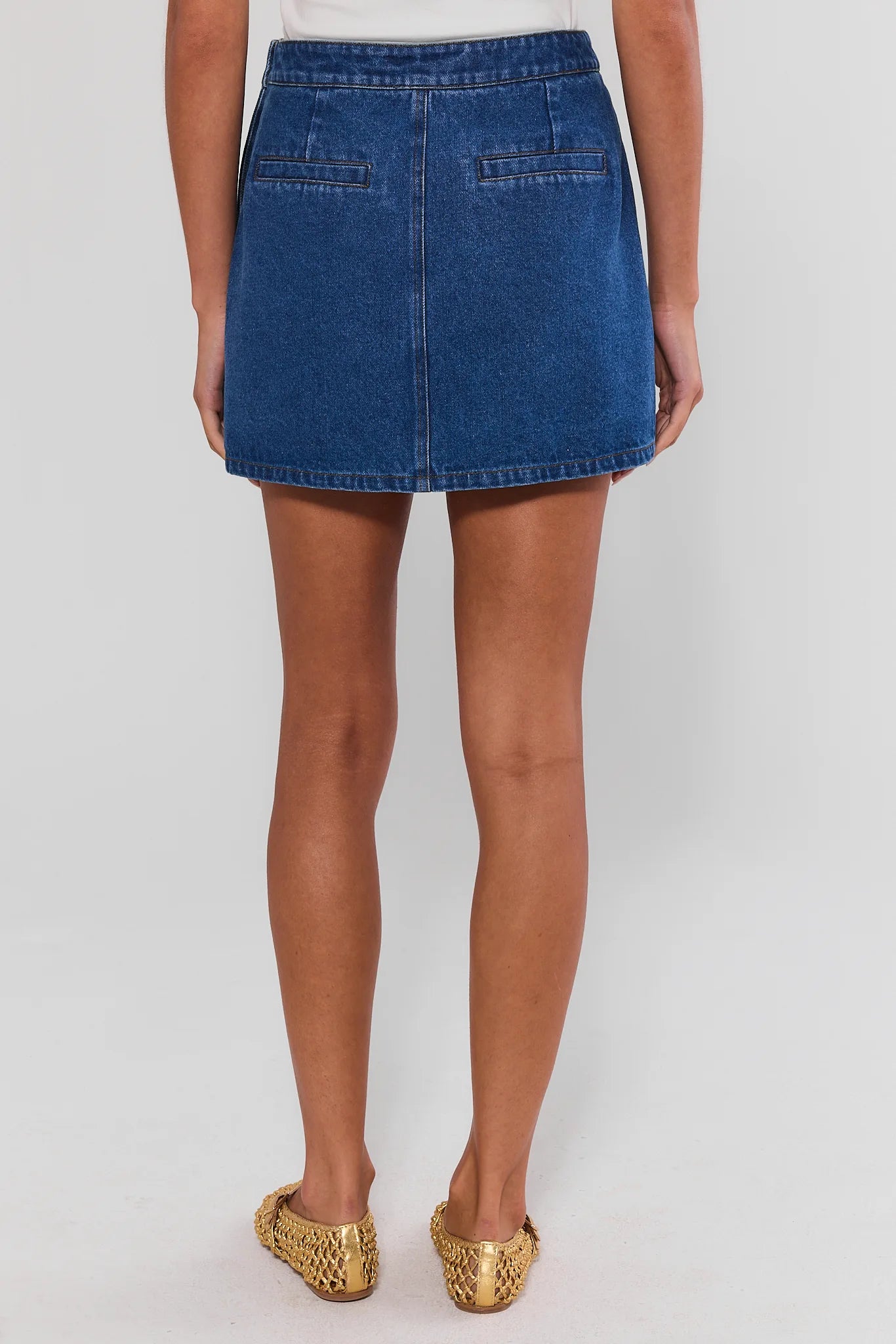 A model shown from the back wearing a blue denim mini skirt, paired with gold snakeskin-patterned flat shoes, against a light gray studio background.