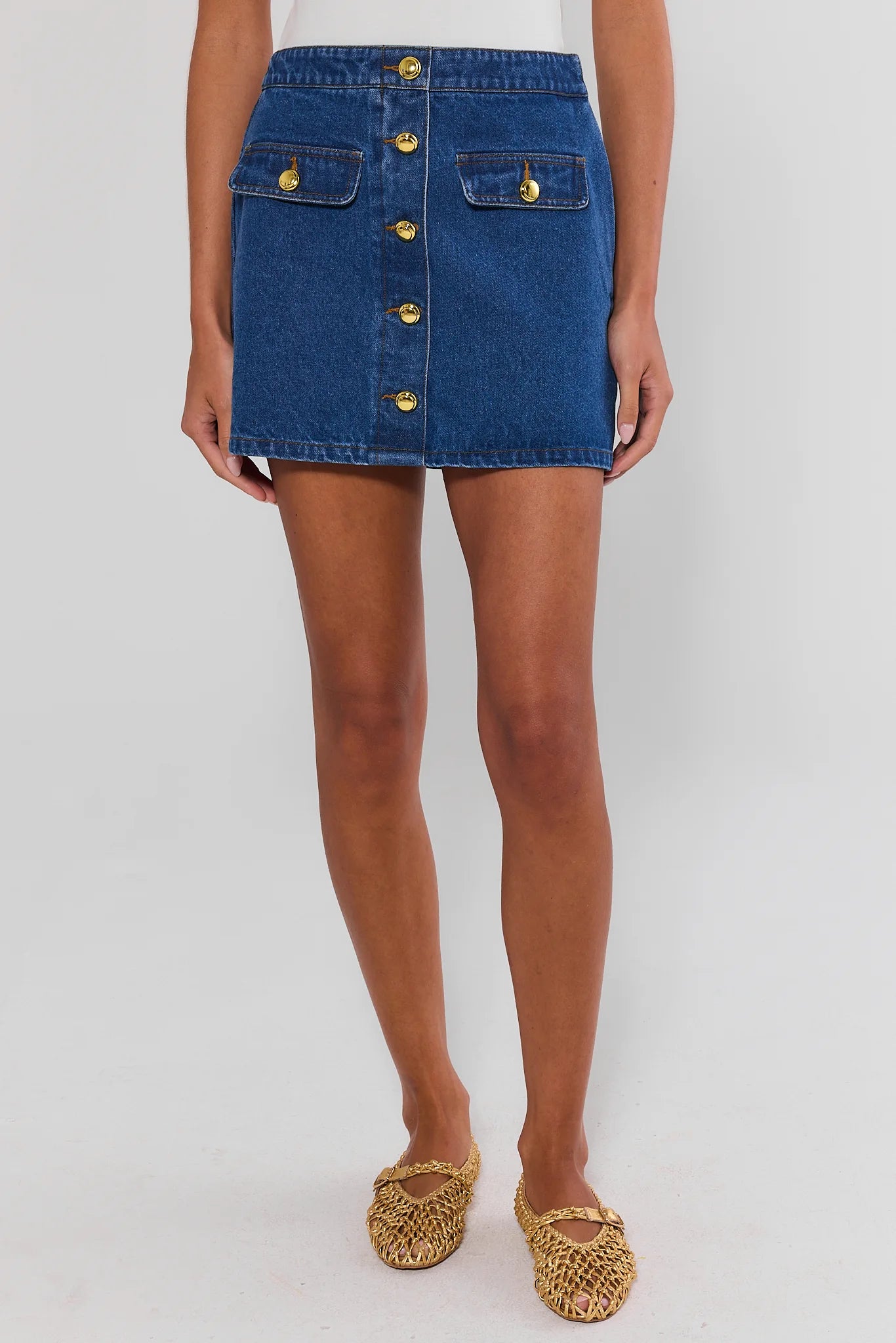 A model wearing a blue denim mini skirt with gold-tone button details and flap pockets, paired with leopard-print flat shoes, against a light gray studio background.
