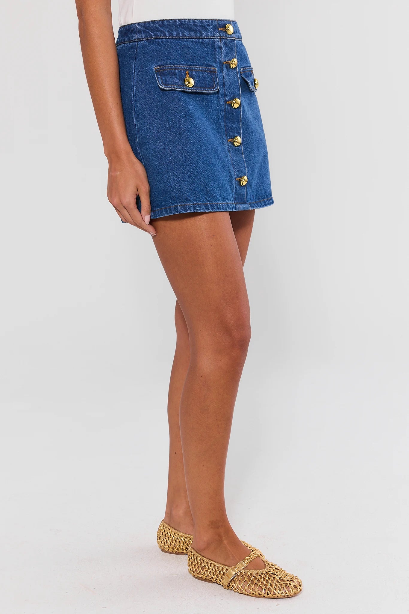 A model wearing a blue denim mini skirt with gold-tone decorative buttons down the front and a flap pocket on the side, paired with woven flat shoes, against a light gray studio background.