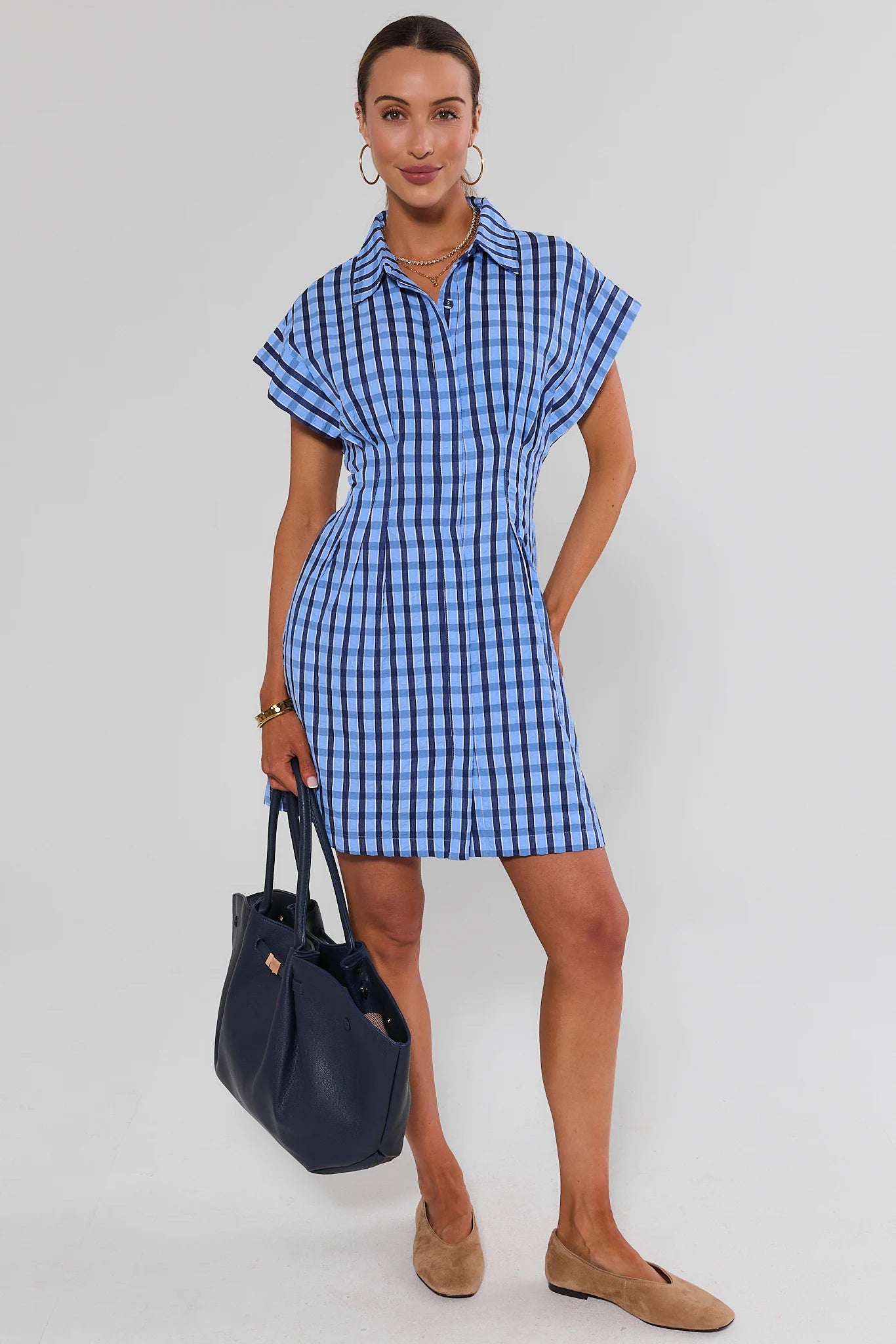 A woman wearing a blue and navy gingham check shirt dress with short flutter sleeves, a pointed collar, and a button-front closure, with flats shown against a light studio background.