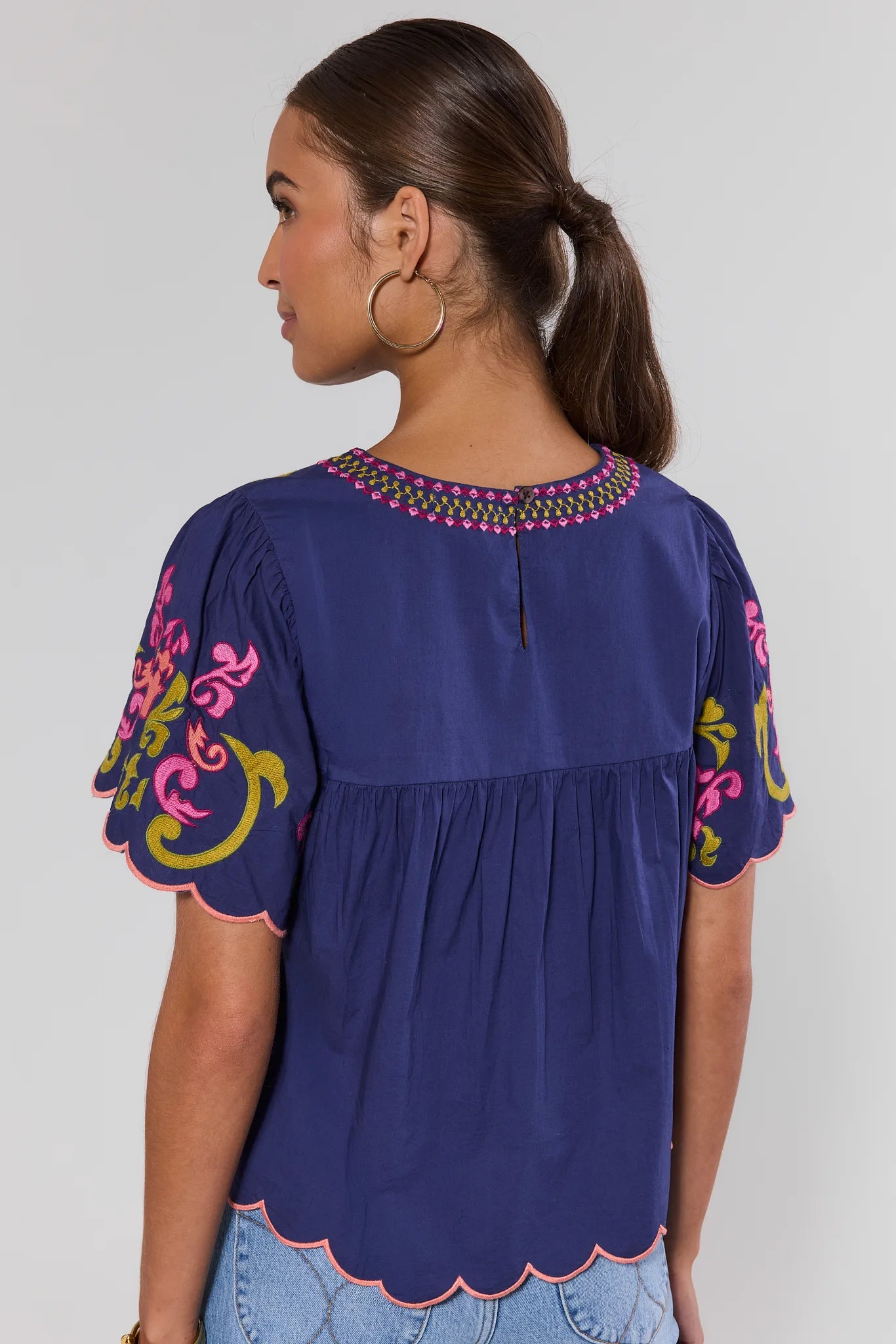 A woman shown from the back wearing a navy blue short-sleeve blouse with colorful embroidery on the sleeves and neckline, paired with light blue jeans against a light gray studio background.