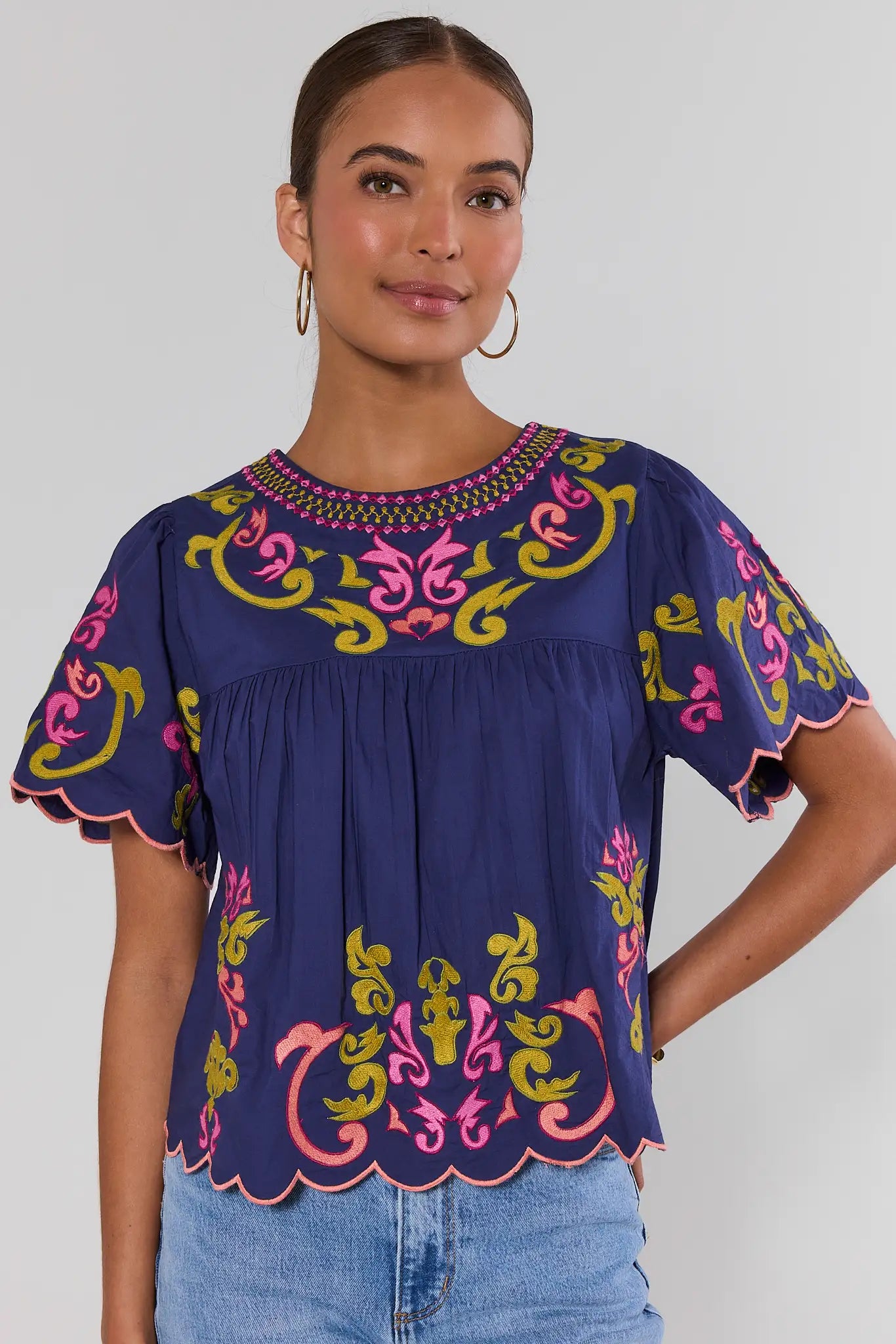 A woman wearing a navy blue blouse with colorful pink, yellow, and green embroidery and scalloped short sleeves, paired with light blue jeans, against a light gray studio background.