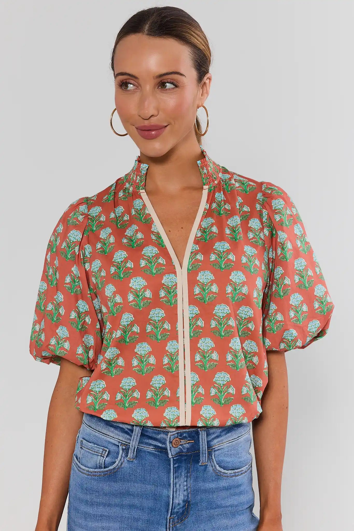 A woman wearing a short-sleeve coral-orange blouse with a green and white floral print, a cream-colored split neckline, and puffed sleeves, paired with blue jeans against a light gray background.