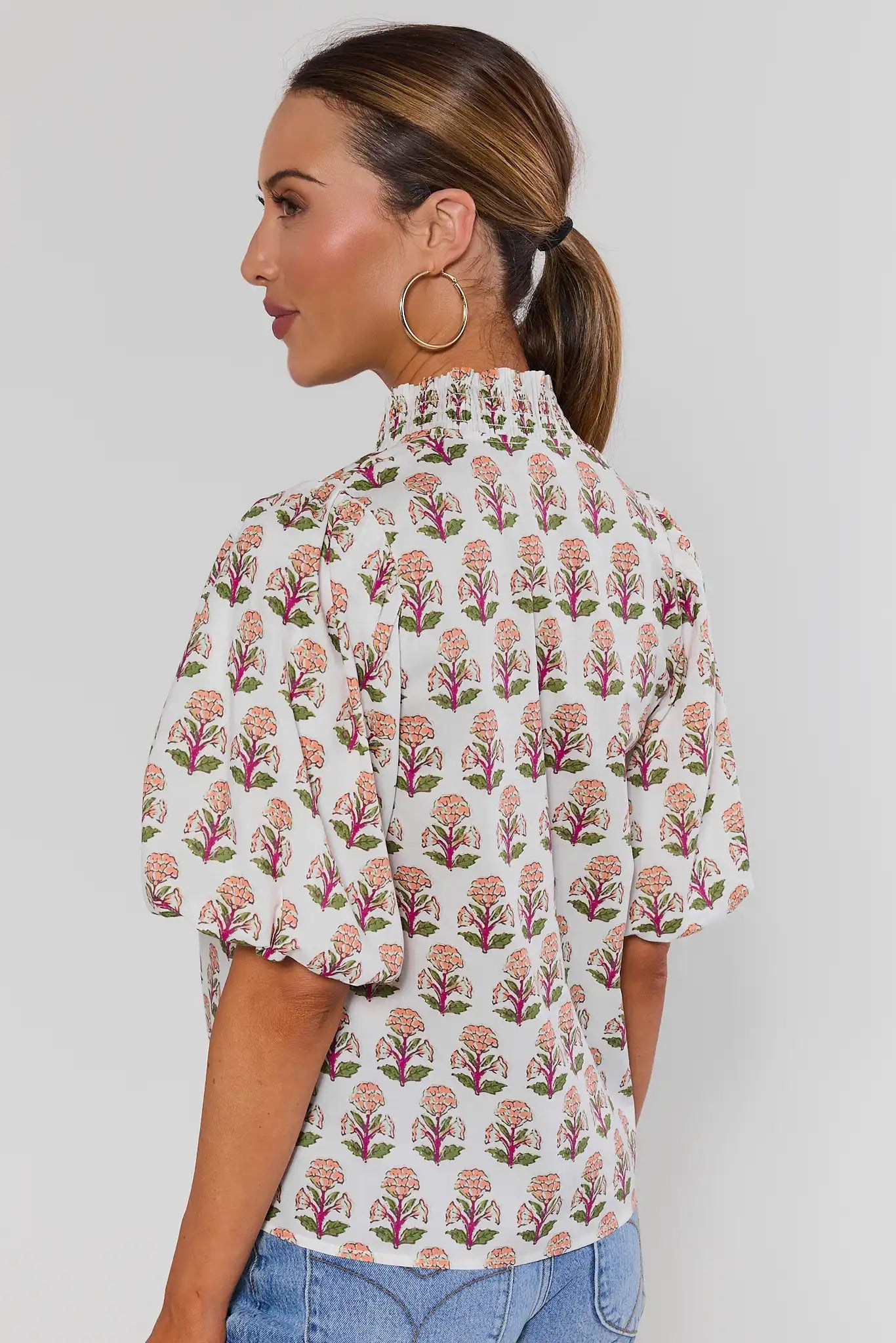 A woman shown from the back wearing a short-sleeve blouse with a white base and an allover pink and green floral block print, paired with blue jeans against a light gray studio background.