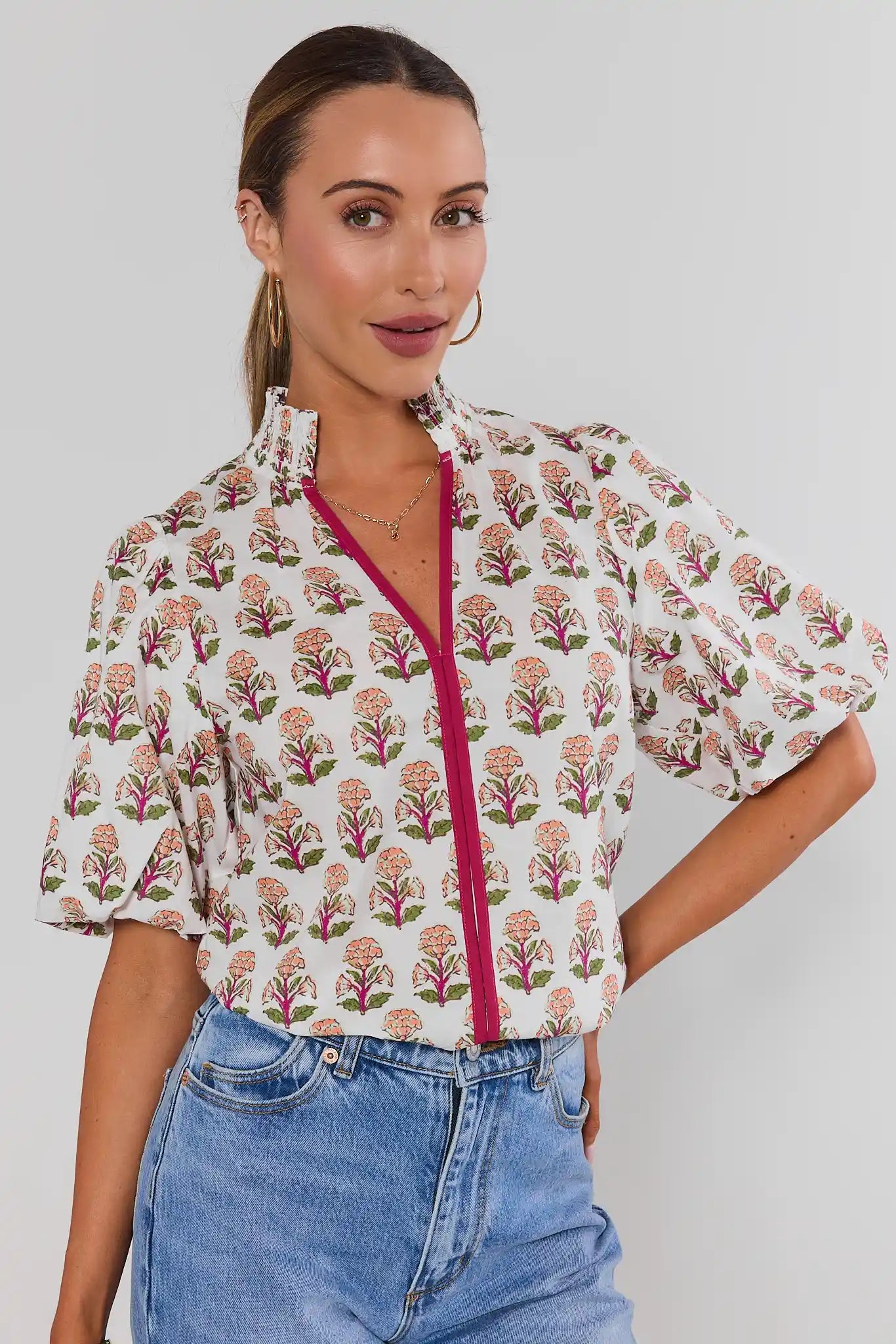 A woman wearing a short-sleeve blouse with a cream background, pink and green floral block print, and a contrasting bright pink front zipper, paired with blue jeans against a light gray background.
