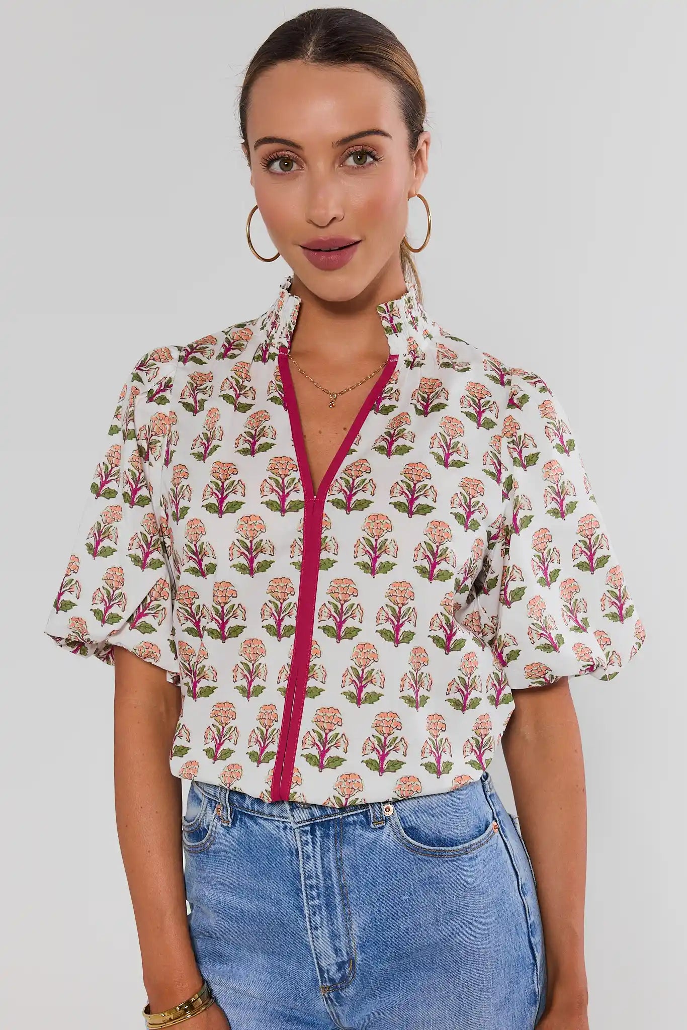 A woman wearing a short-sleeve button-up blouse with a white base, pink and green floral block print, and a contrasting bright pink front zipper, paired with blue jeans against a light gray studio background.
