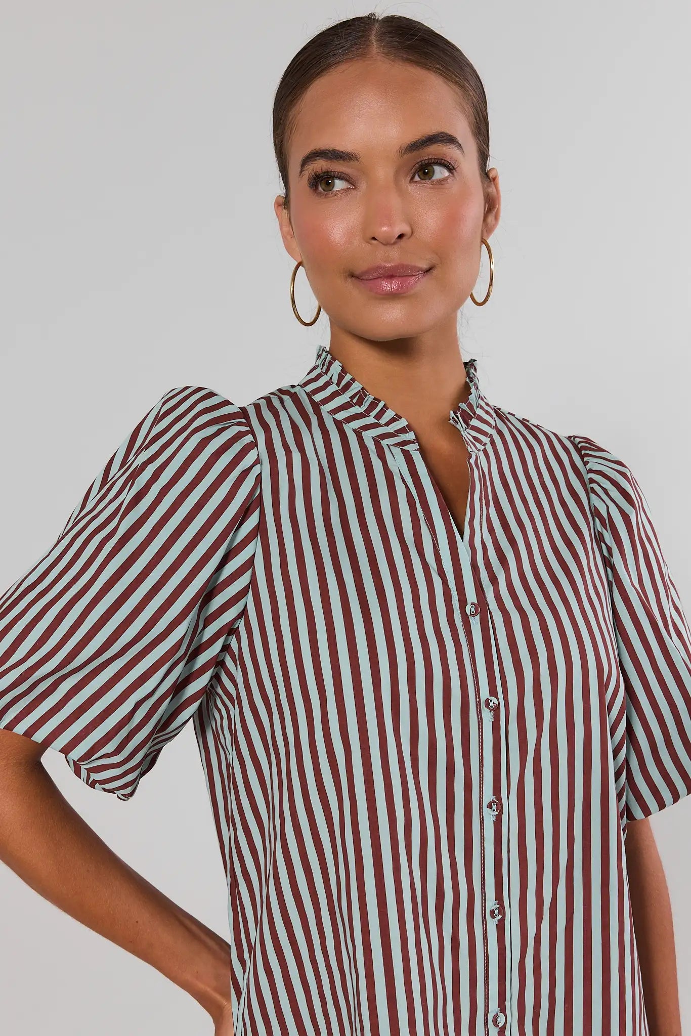 A woman wearing a short-sleeve button-front blouse with vertical burgundy and mint green stripes, paired with gold hoop earrings, against a plain light gray studio background.