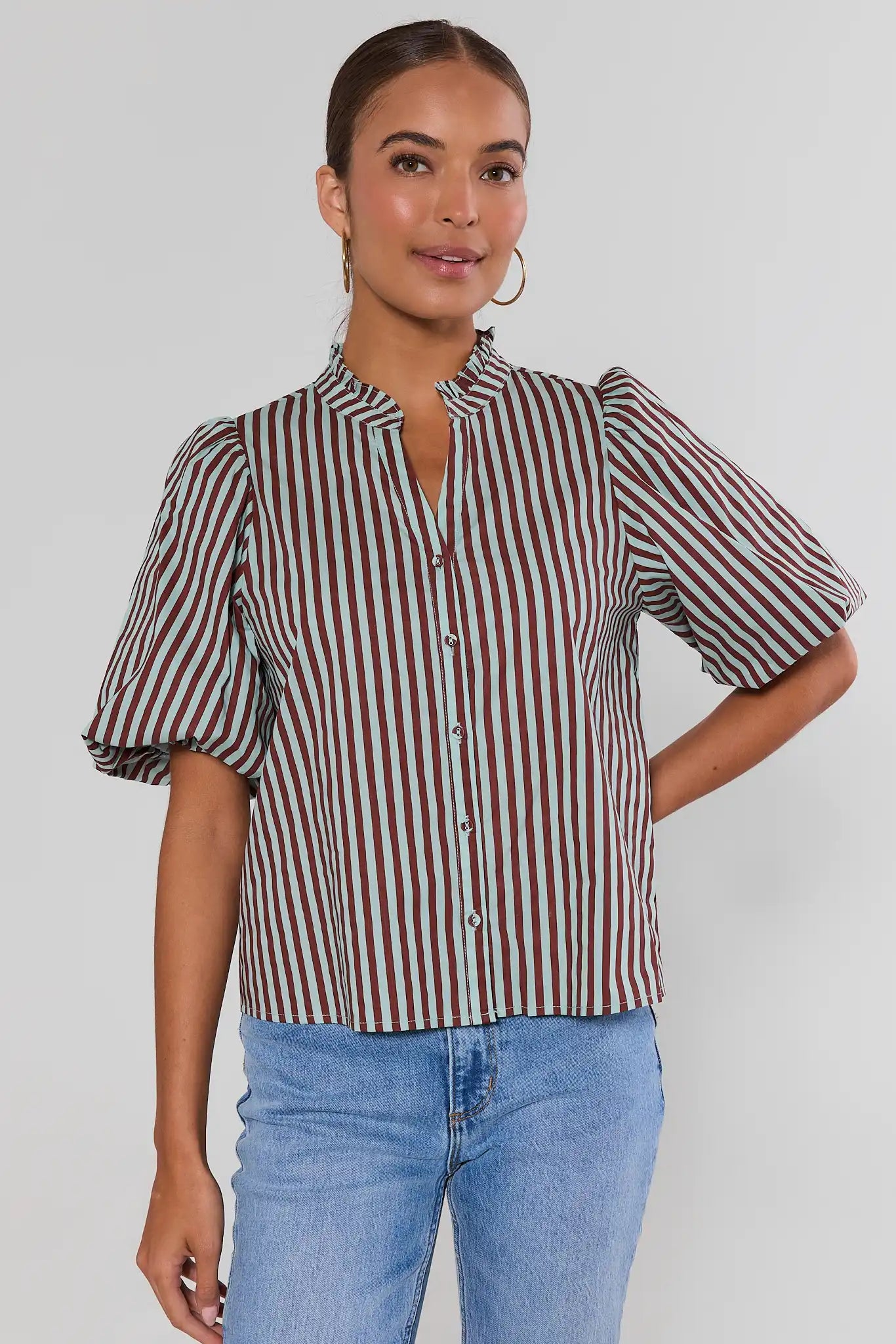 A woman wearing a short-sleeve button-front blouse with vertical burgundy and mint green stripes, paired with gold hoop earrings, against a plain light background.