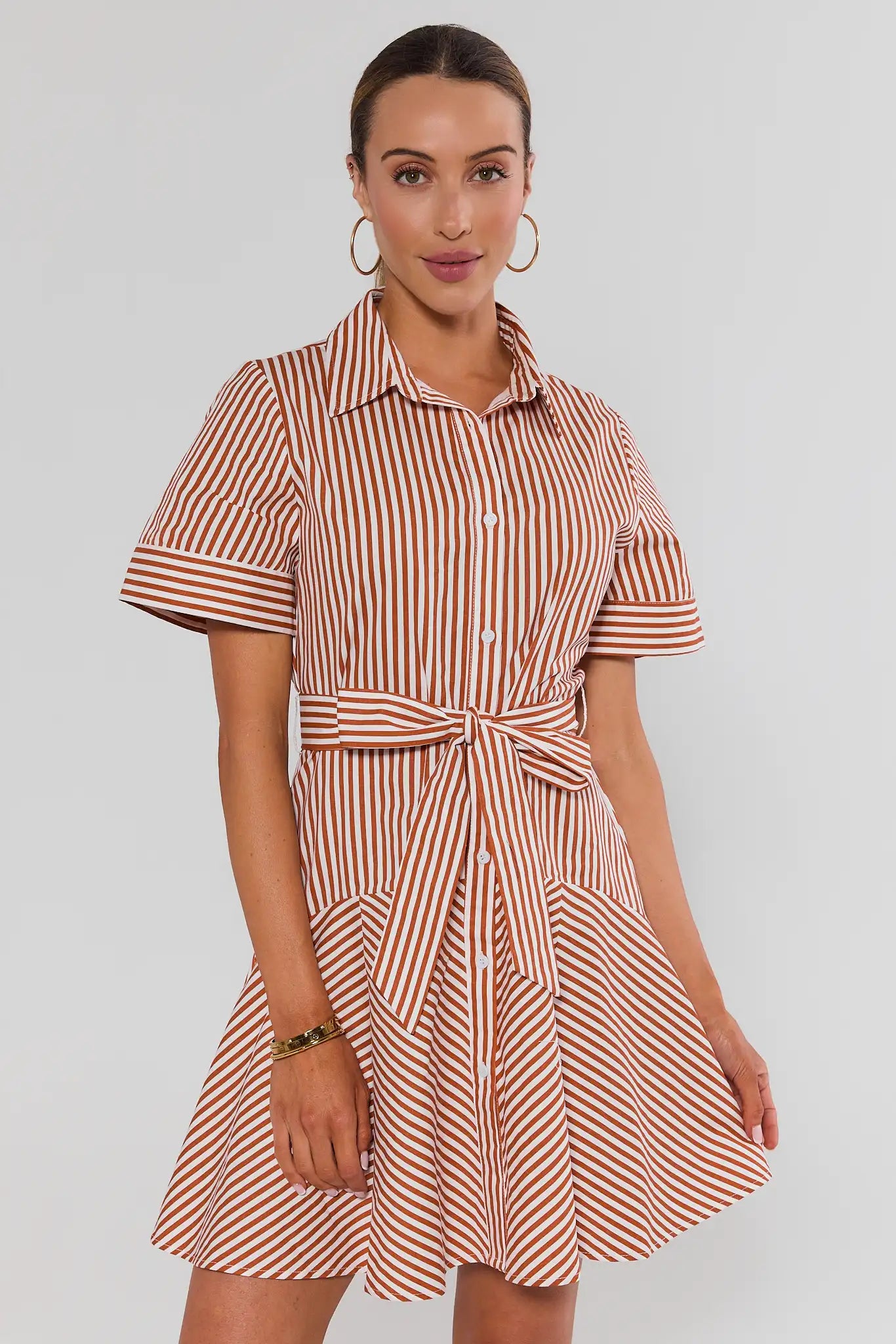 A woman modeling a short-sleeve collared shirt dress with vertical red and white stripes, featuring a tie waist and flared skirt, against a light gray studio background.