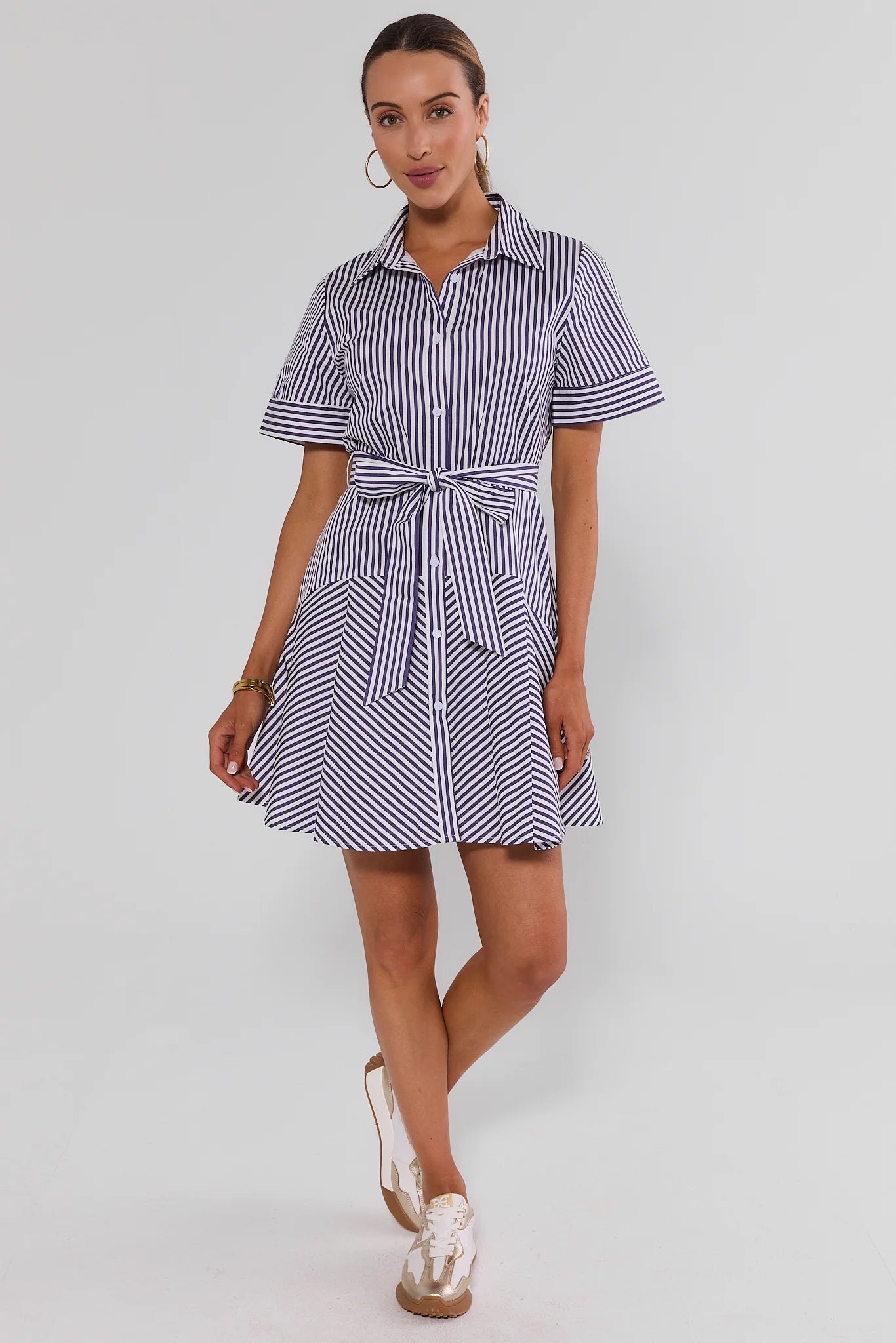 A woman walking wearing a short-sleeve collared shirt dress with vertical navy and white stripes, featuring a tie waist belt, photographed against a light studio background.
