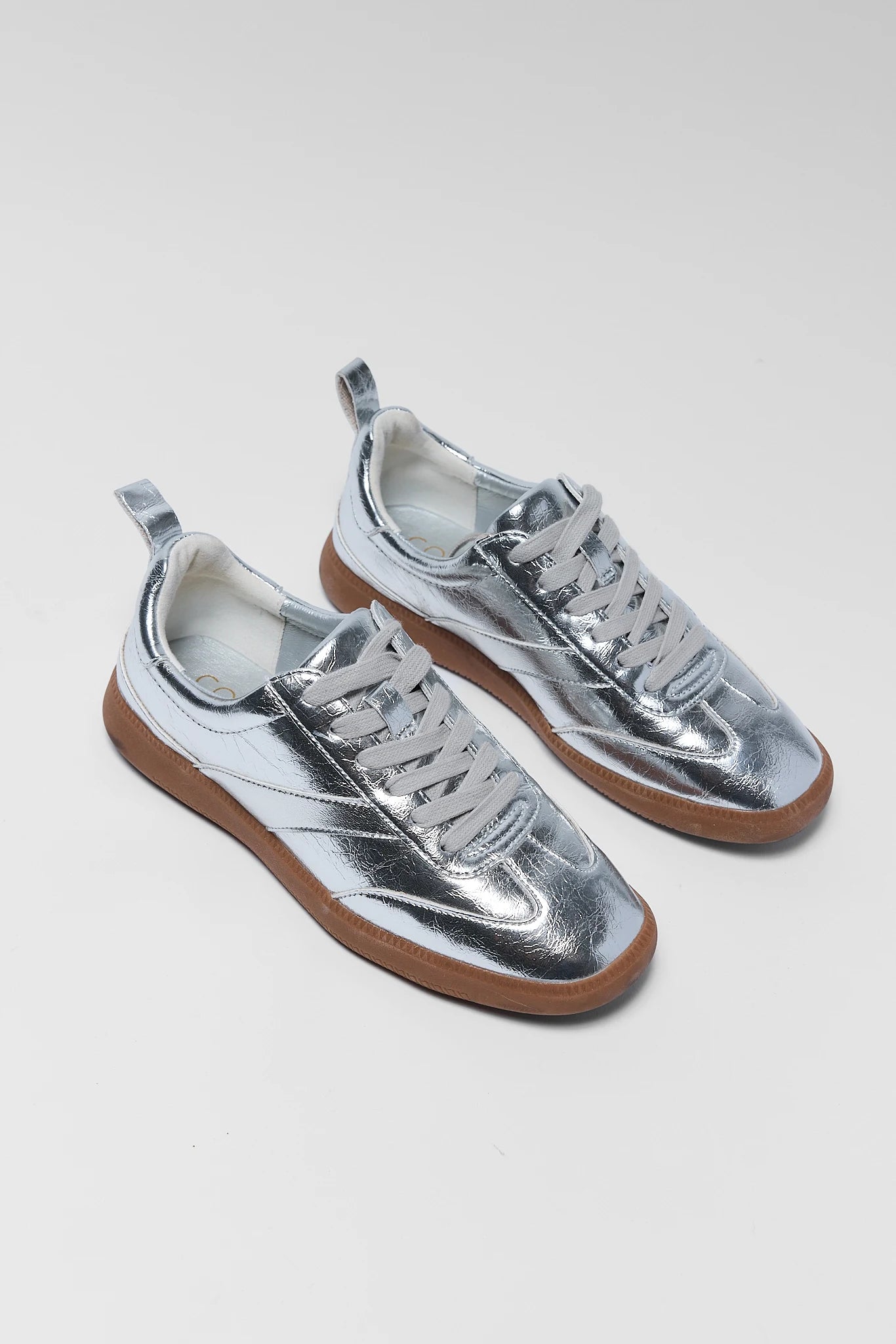 A pair of metallic silver low-top sneakers with brown rubber soles, displayed on a light gray background.