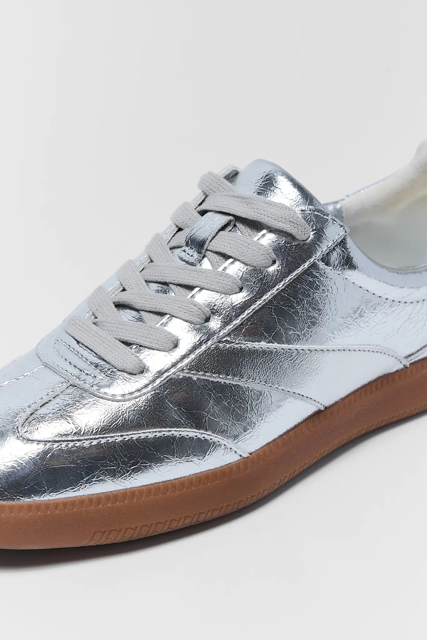 A close-up of a metallic silver low-top sneaker with gray laces and a brown gum sole, shown against a light gray background.