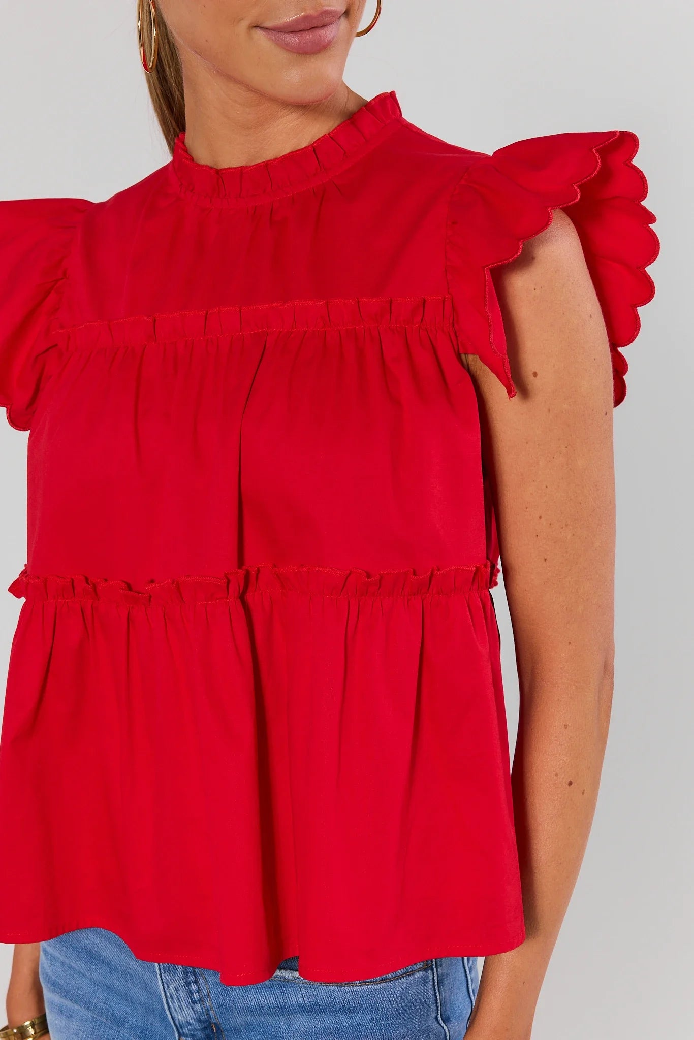 Close up a woman modeling a bright red ruffled short-sleeve top with a high neckline, against a light gray studio background.