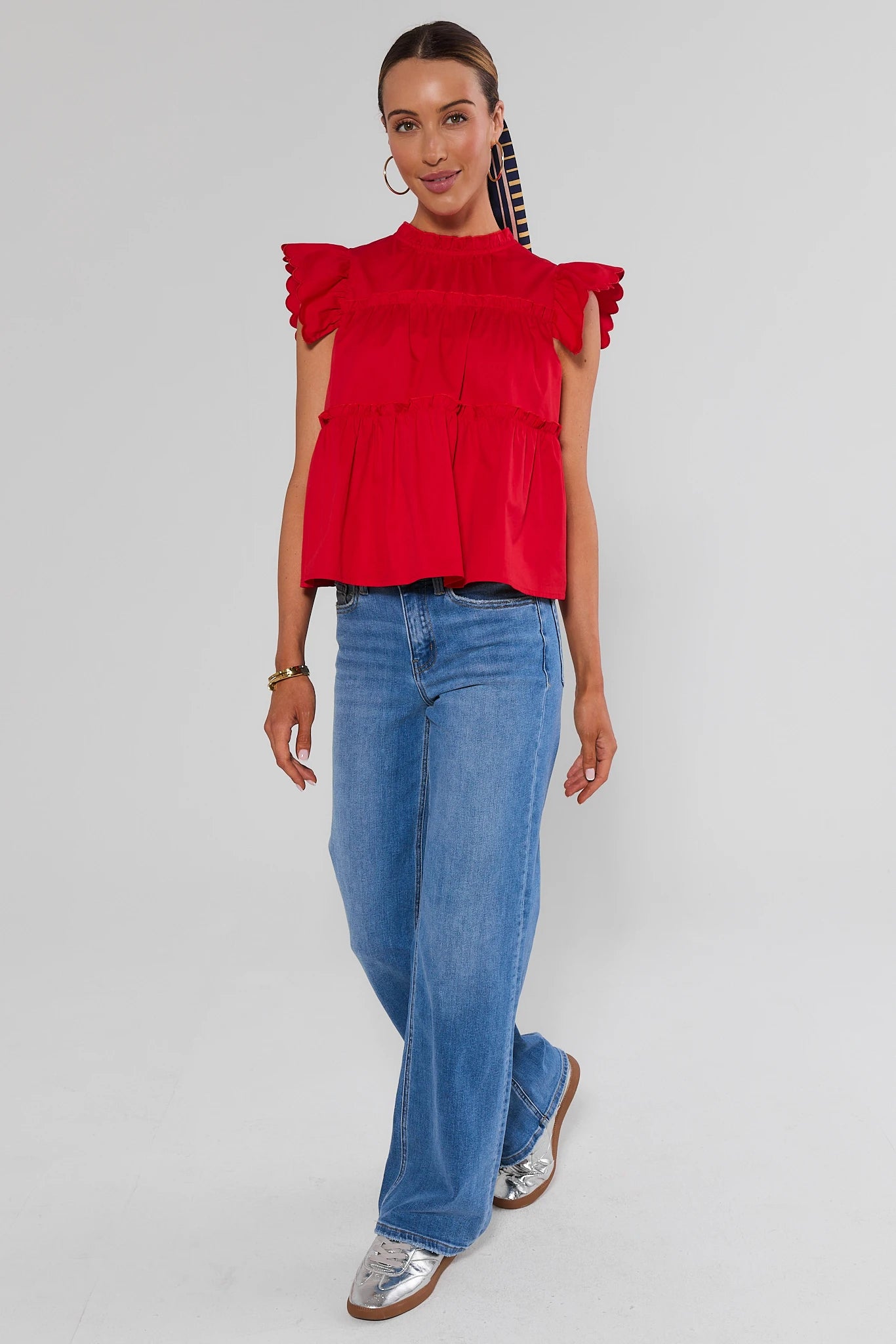 A woman modeling a bright red ruffled short-sleeve top with a high neckline, paired with wide-leg blue jeans and silver loafers, against a light gray studio background.