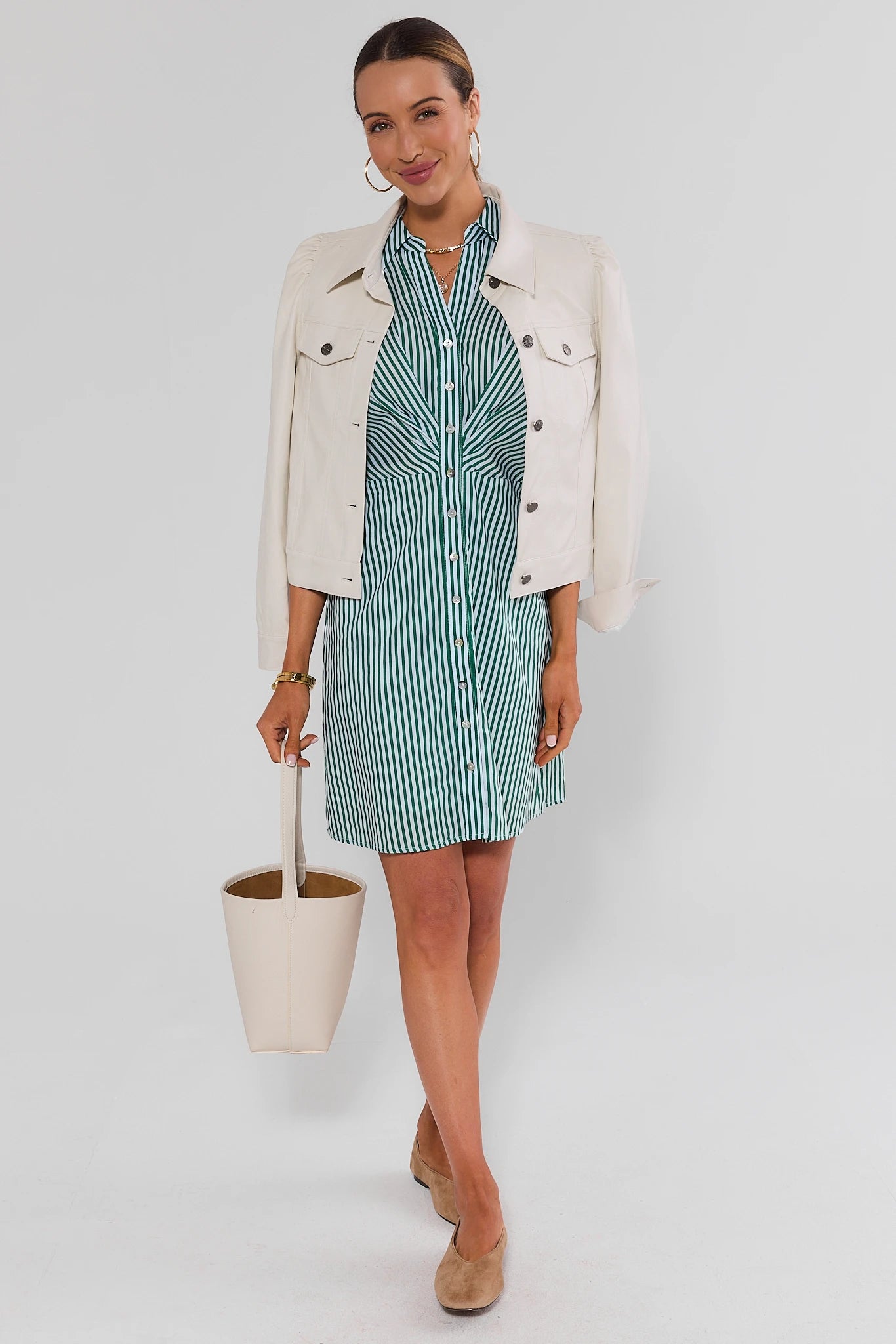 A woman models a green and white striped shirt dress with a matching tie-neck detail, styled with a cream-colored denim jacket, a cream bucket bag, and tan suede loafers, against a light gray studio background.