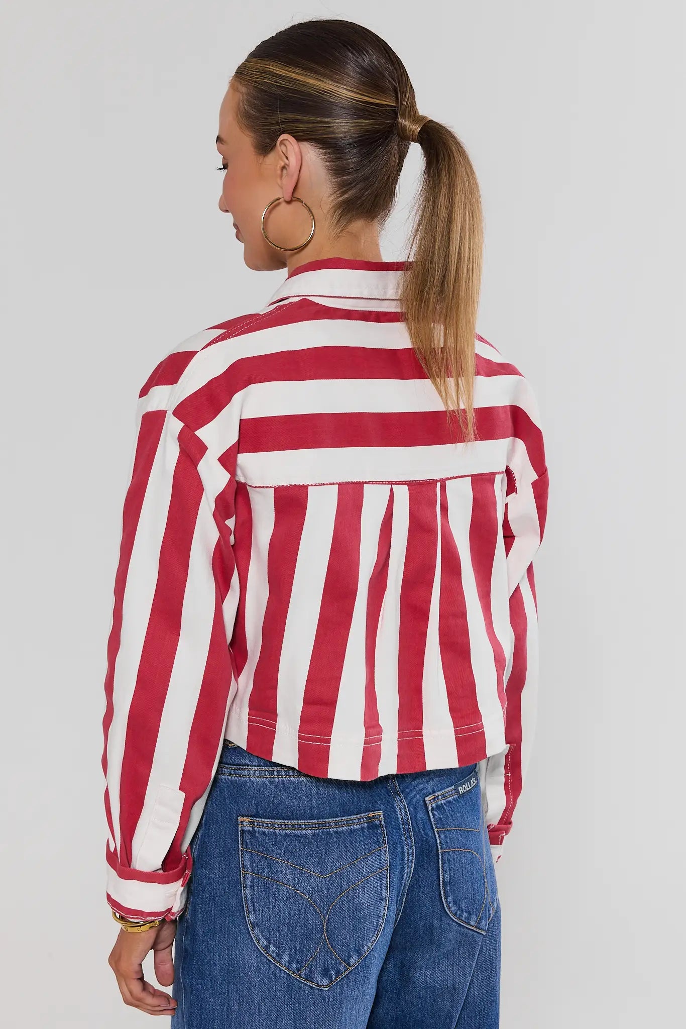 A woman shown from the back wearing a red and white vertically striped cropped jacket over a white top and blue jeans, against a light gray studio background.