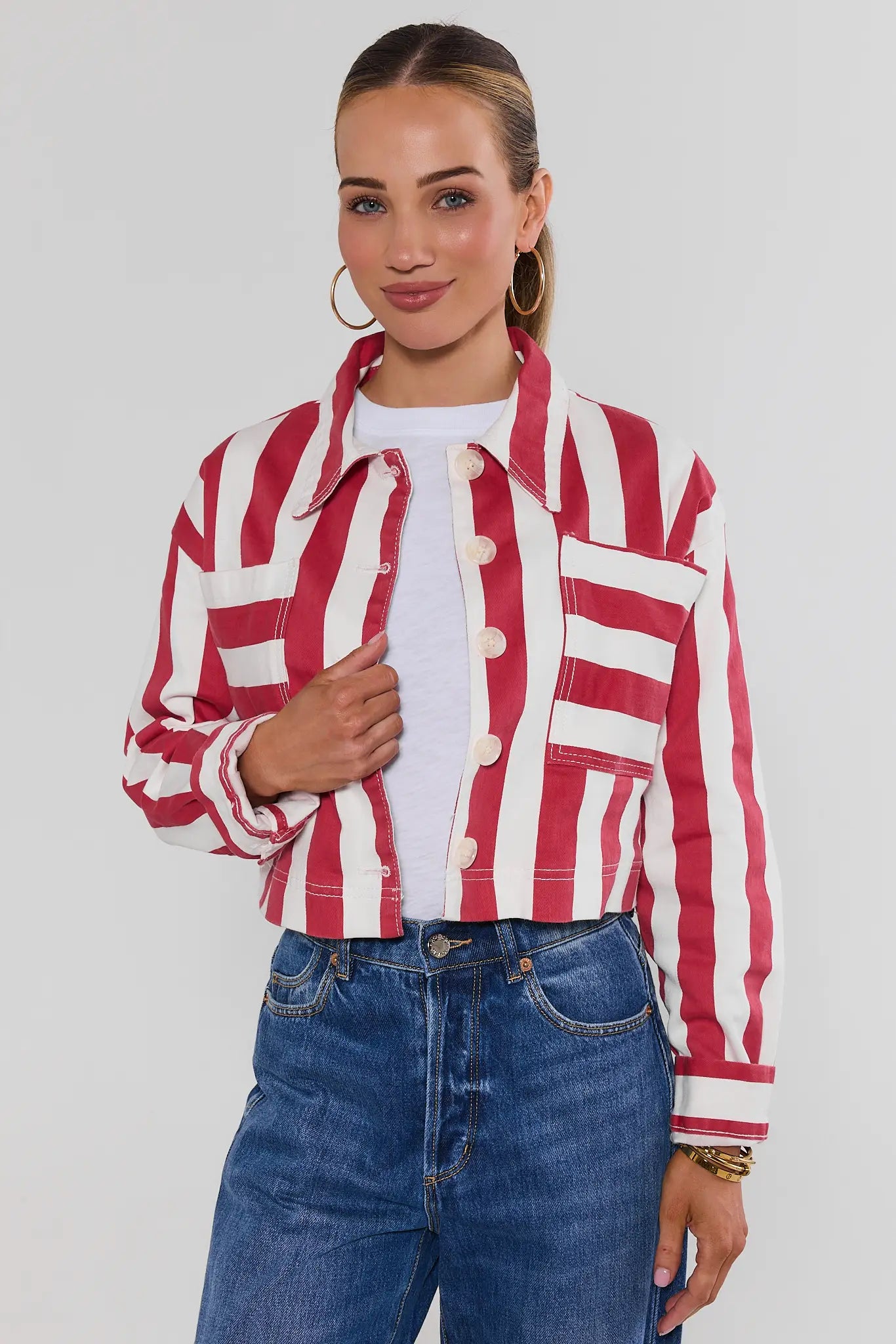 A woman modeling a cropped red and white striped button-up jacket over a white t-shirt and blue jeans, set against a plain light gray studio background.
