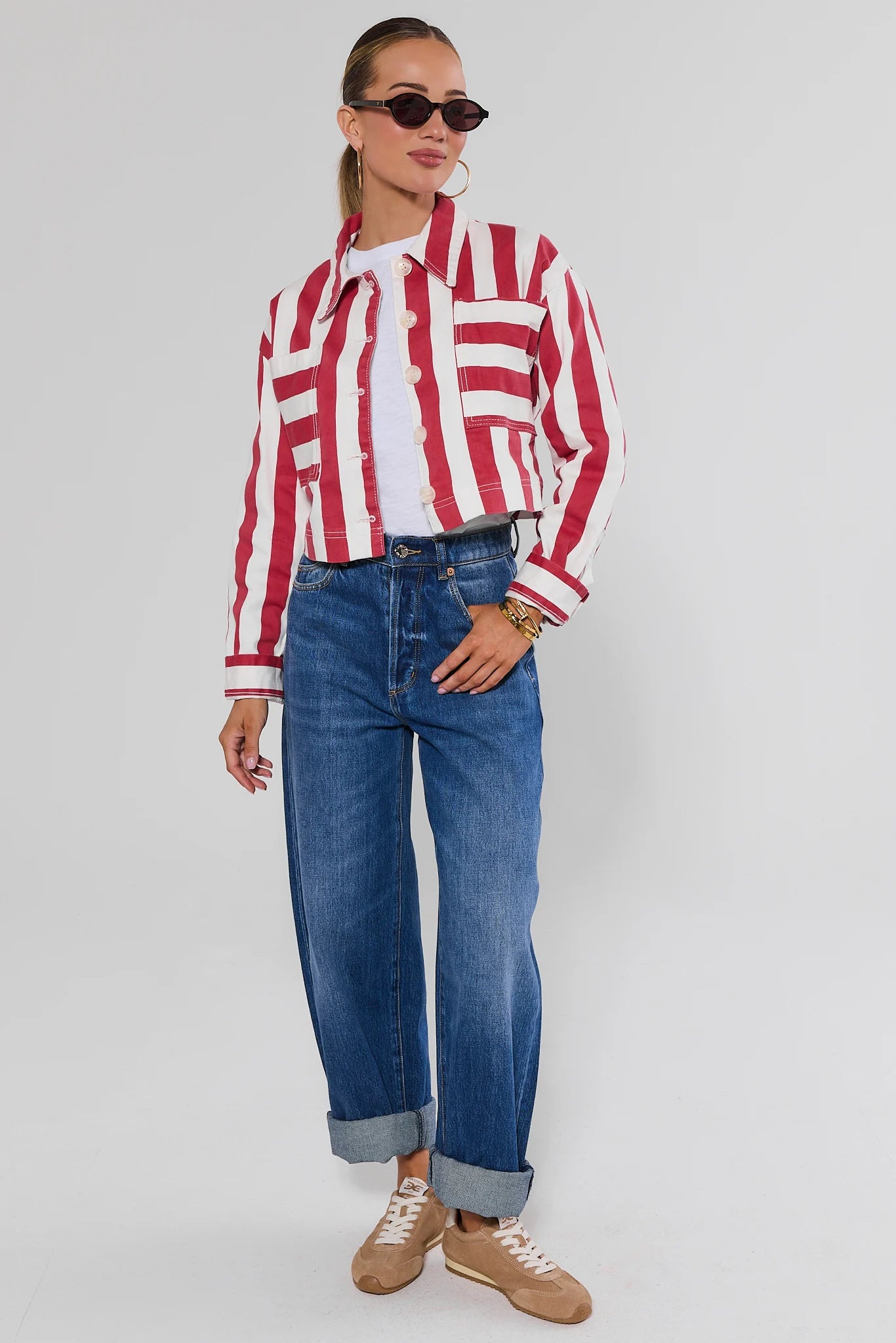 A woman modeling a red and white vertically striped button-up shirt, blue wide-leg jeans, and tan sneakers against a light gray studio background.